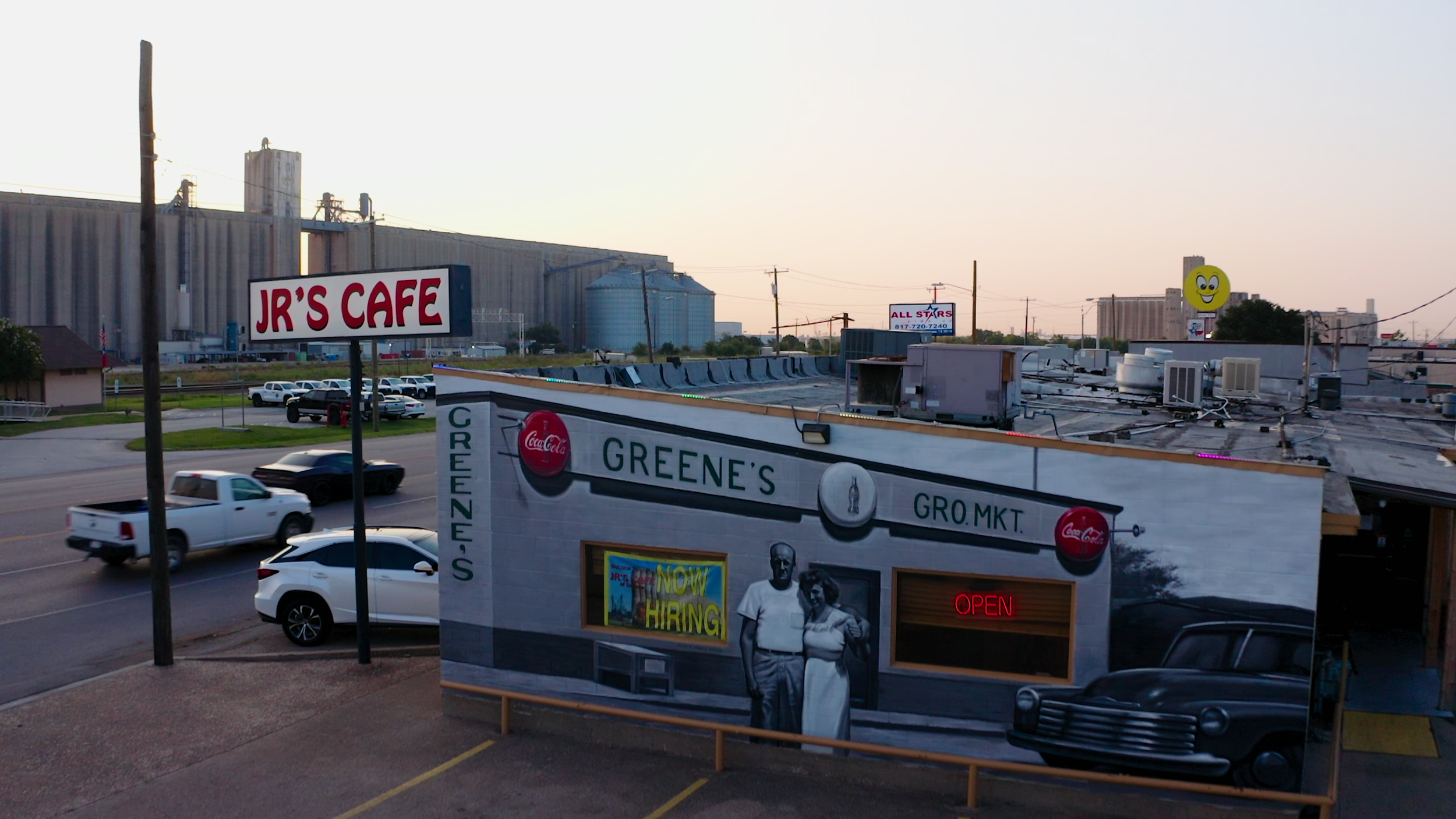 Street view of a small business with a painted facade, a neon 'Open' sign, and a large sign for JR's Cafe in the background. Several cars are parked and driving by on the street.