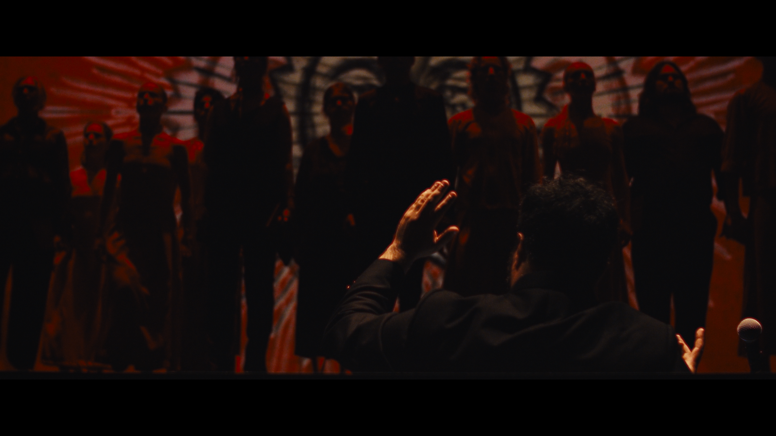 A person conducts a choir of nine people on stage, with a background displaying a dark, shadowy, and patterned projection. The conductor, with one hand raised, faces the choir members who are illuminated faintly and wearing dark clothing, and some ar