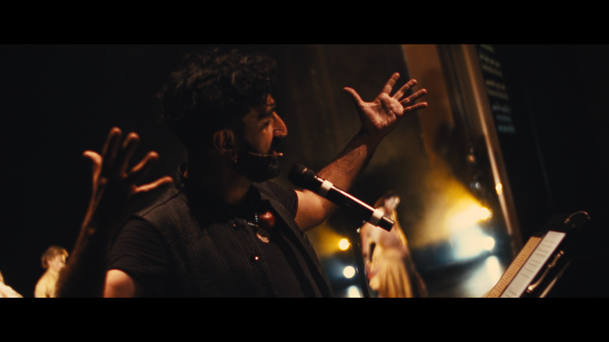 A man with curly hair and a beard speaking into a microphone on stage, raising his hand with fingers spread, with a blurred background and stage lighting.