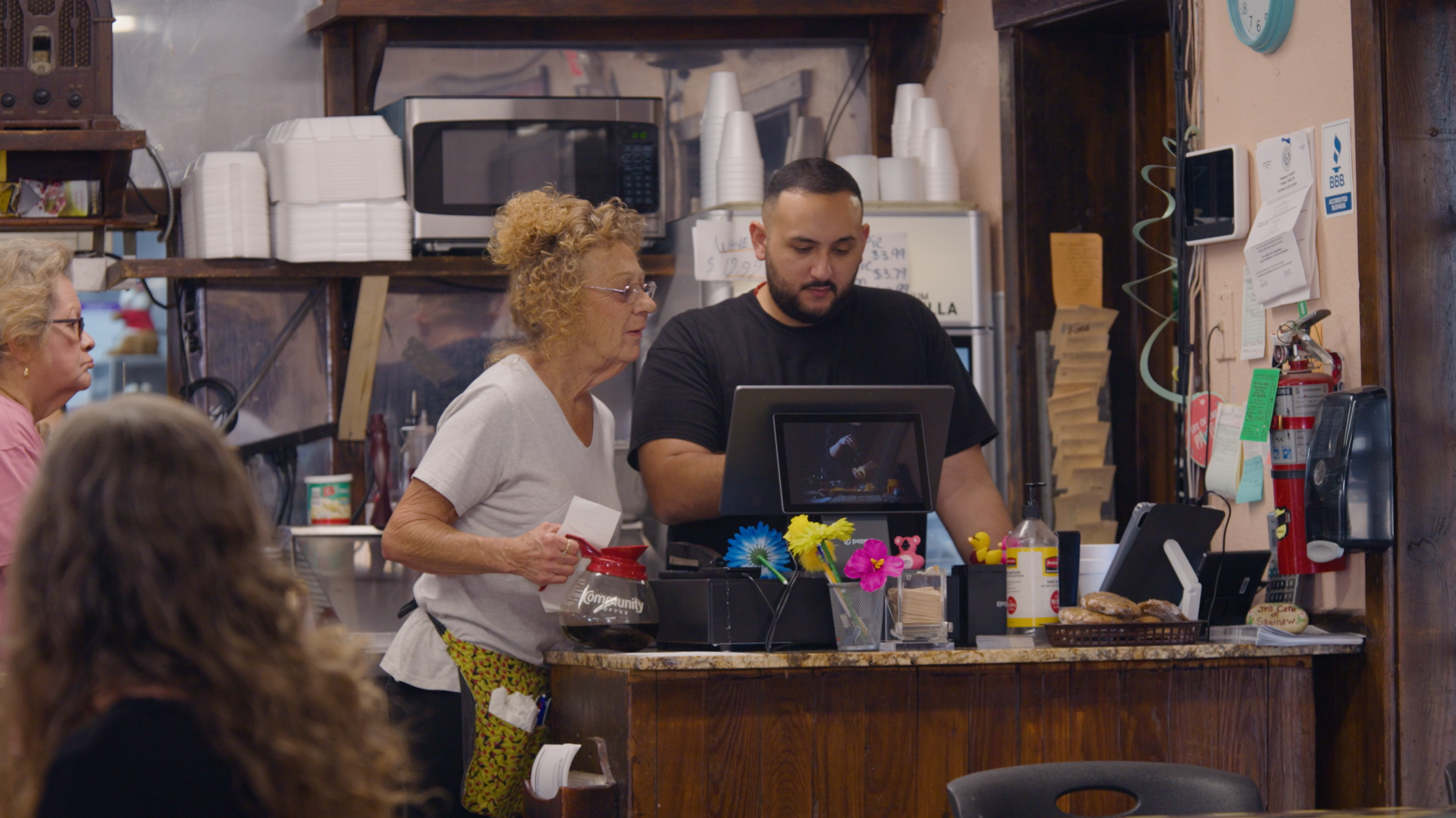 A woman with curly blond hair and glasses is ordering at a counter in a casual restaurant while a young man with a beard and black shirt is assisting her with a point-of-sale system. There are several older women waiting nearby, and the background in