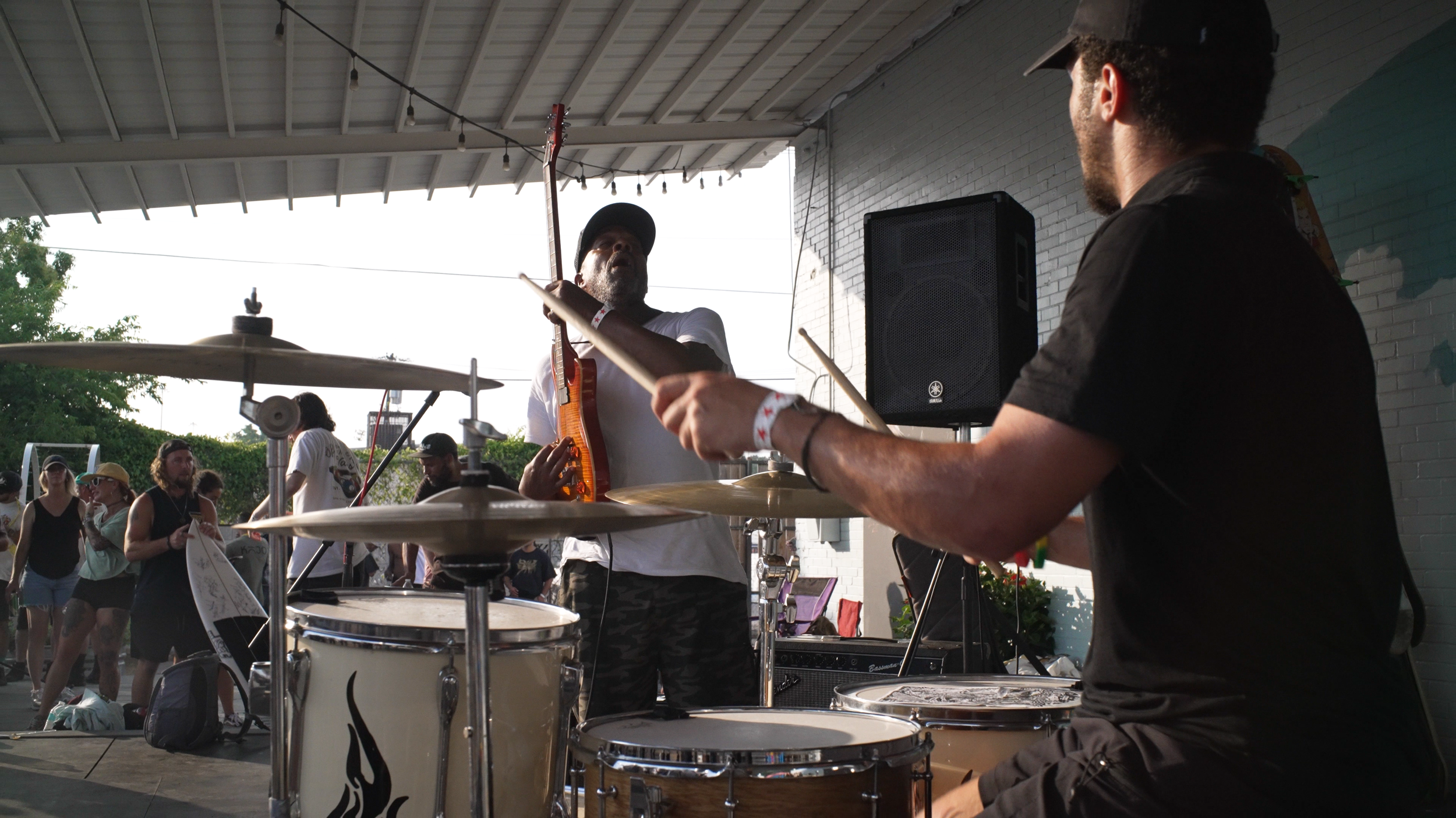 Musicians perform at an outdoor event, with a drummer and guitarist on stage, and a crowd of people in the background.