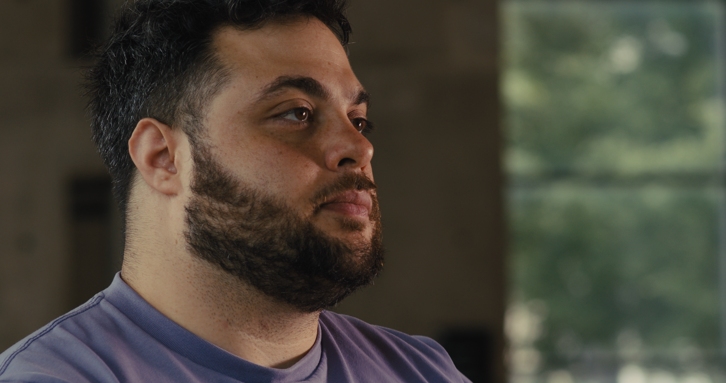 Close-up of a young man with dark hair and a beard, wearing a purple shirt, looking thoughtfully to the side indoors near a window with blurred greenery outside.