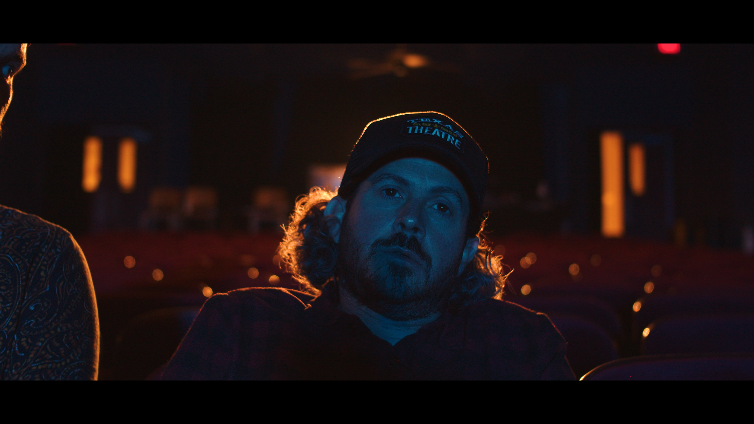 A man with long hair and a beard, wearing a black cap with a theatre logo, sitting in a dimly lit theatre or cinema with red seats, looking thoughtful or focused.