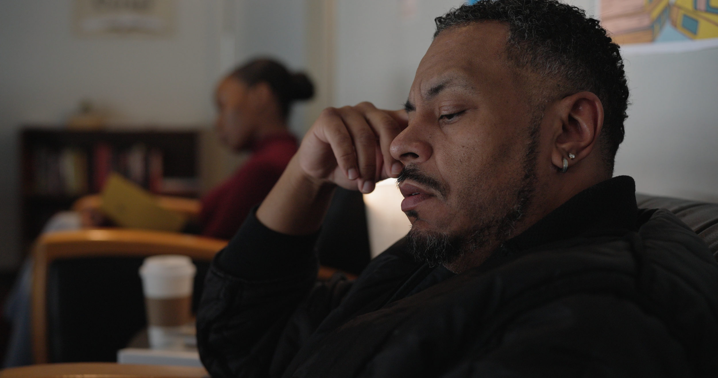 A man with short curly hair and earrings sitting with his hand touching his face, appearing upset or deep in thought, in a setting with a blurred woman reading a book in the background.