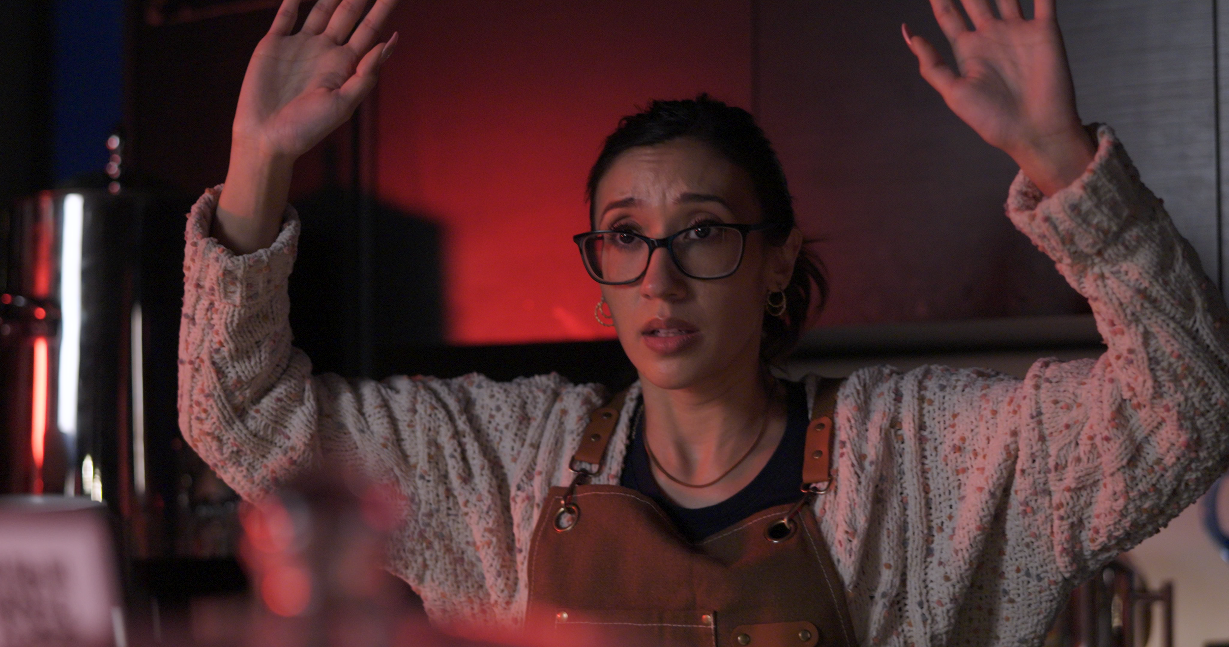 A woman with glasses and dark hair, wearing a knitted sweater and apron, raises her hands with a worried or surprised expression in a dimly lit kitchen.