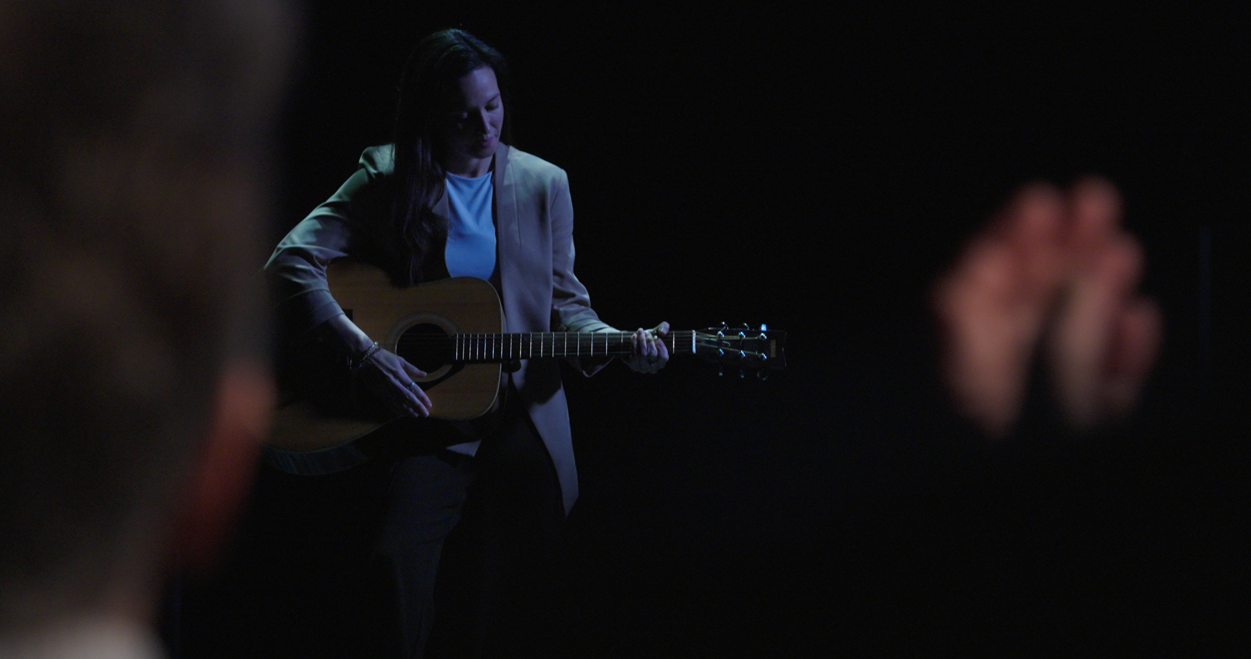 A woman is playing an acoustic guitar on a dark stage.