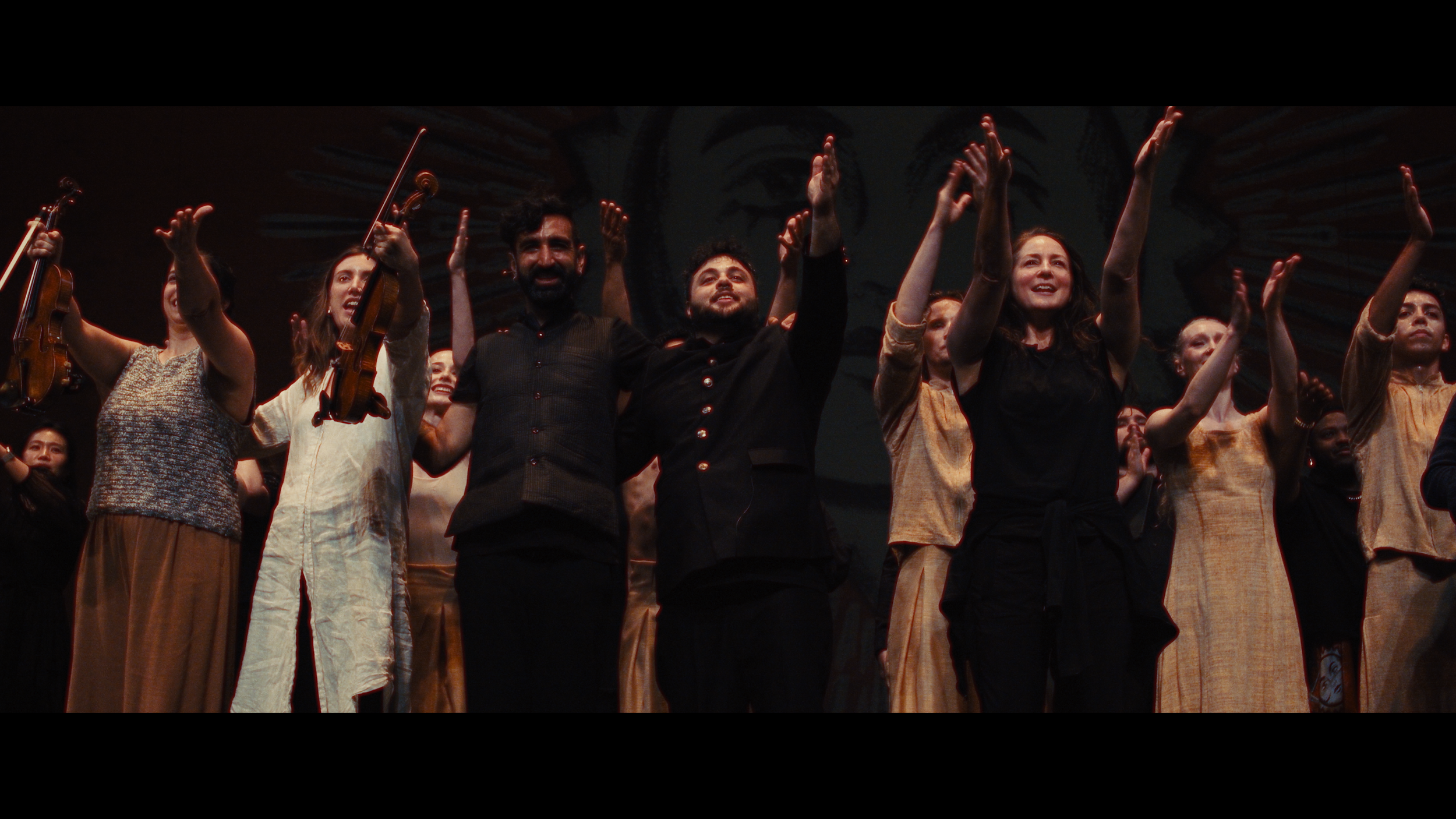 Group of performers on stage, some holding violins, taking a bow or concertmaster, clapping, and smiling.