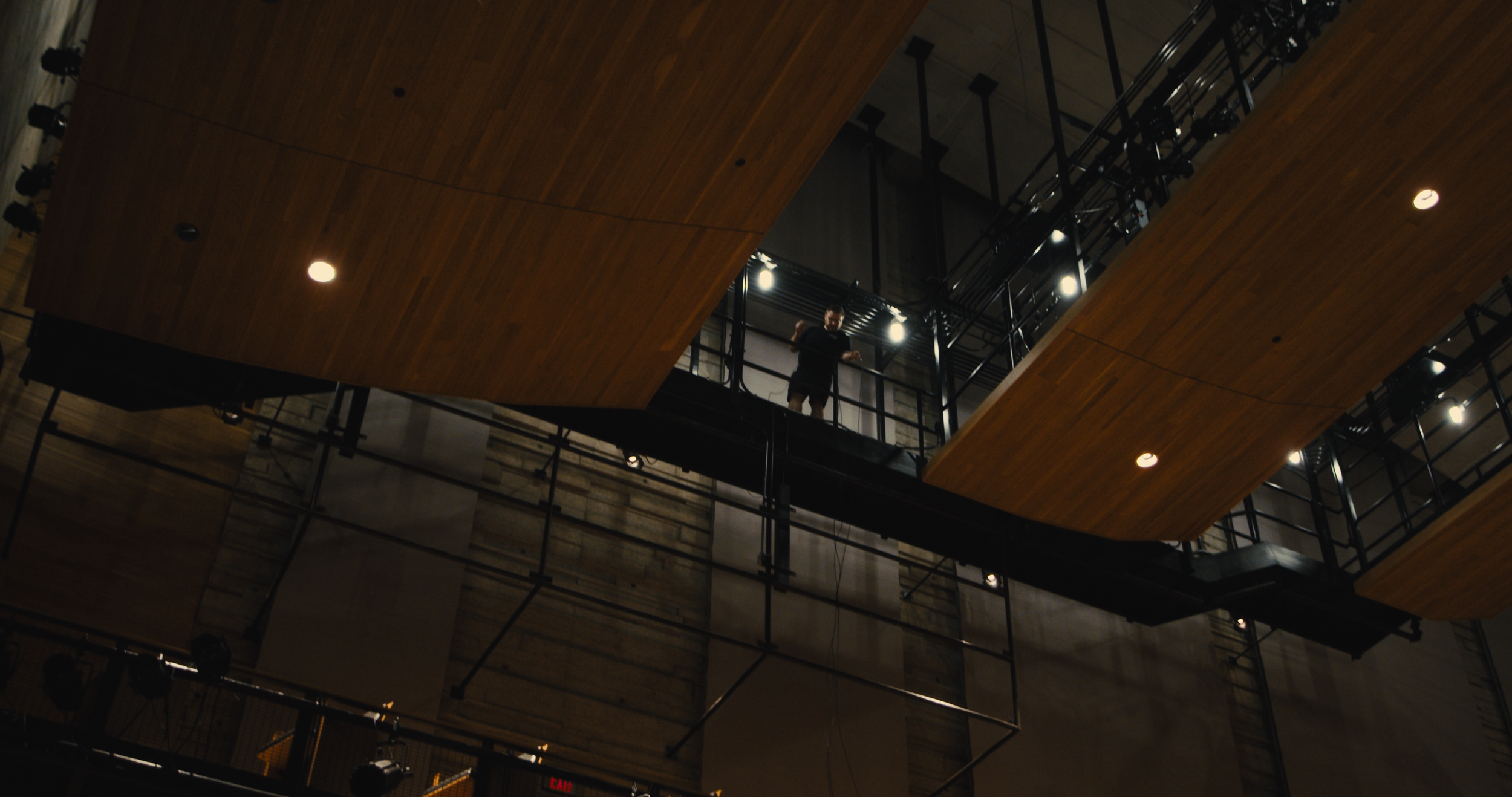 A person standing on an upper balcony of a theater or concert hall, looking down, with wooden panels and stage lighting visible.