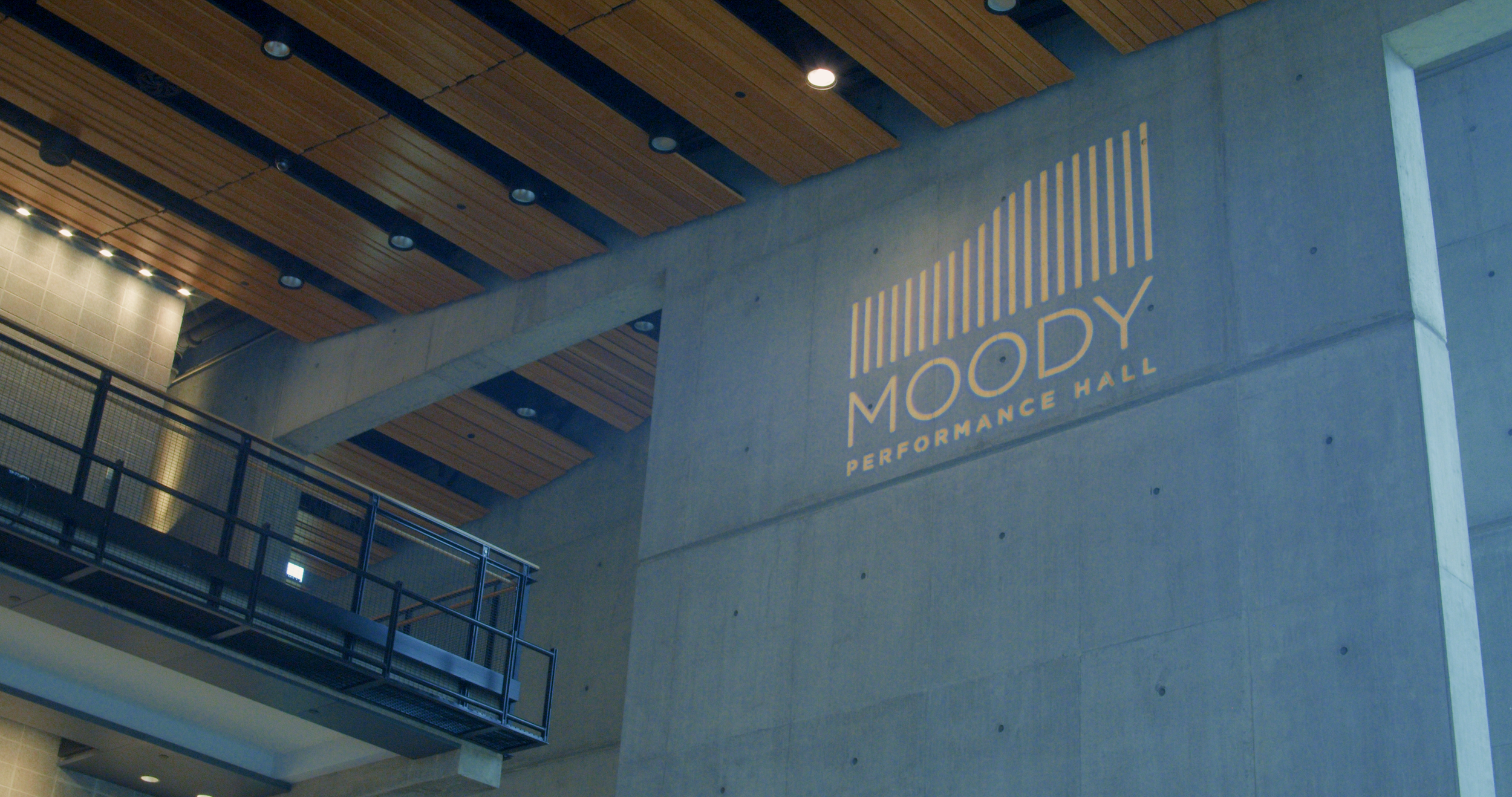 Interior view of a performance hall named Moody Performance Hall with a large concrete wall, a sign with the hall's name, wooden ceiling panels, and a balcony with metal railings.