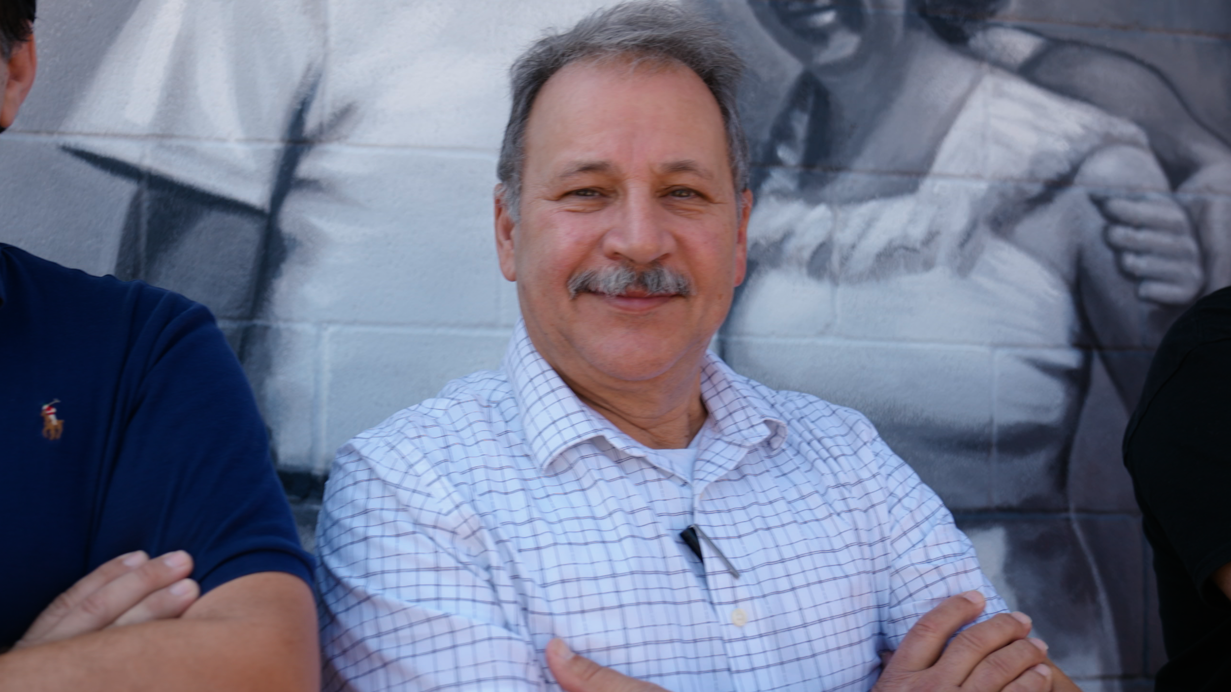 A smiling middle-aged man with gray hair and a mustache standing with arms crossed in front of a mural of a person fishing.