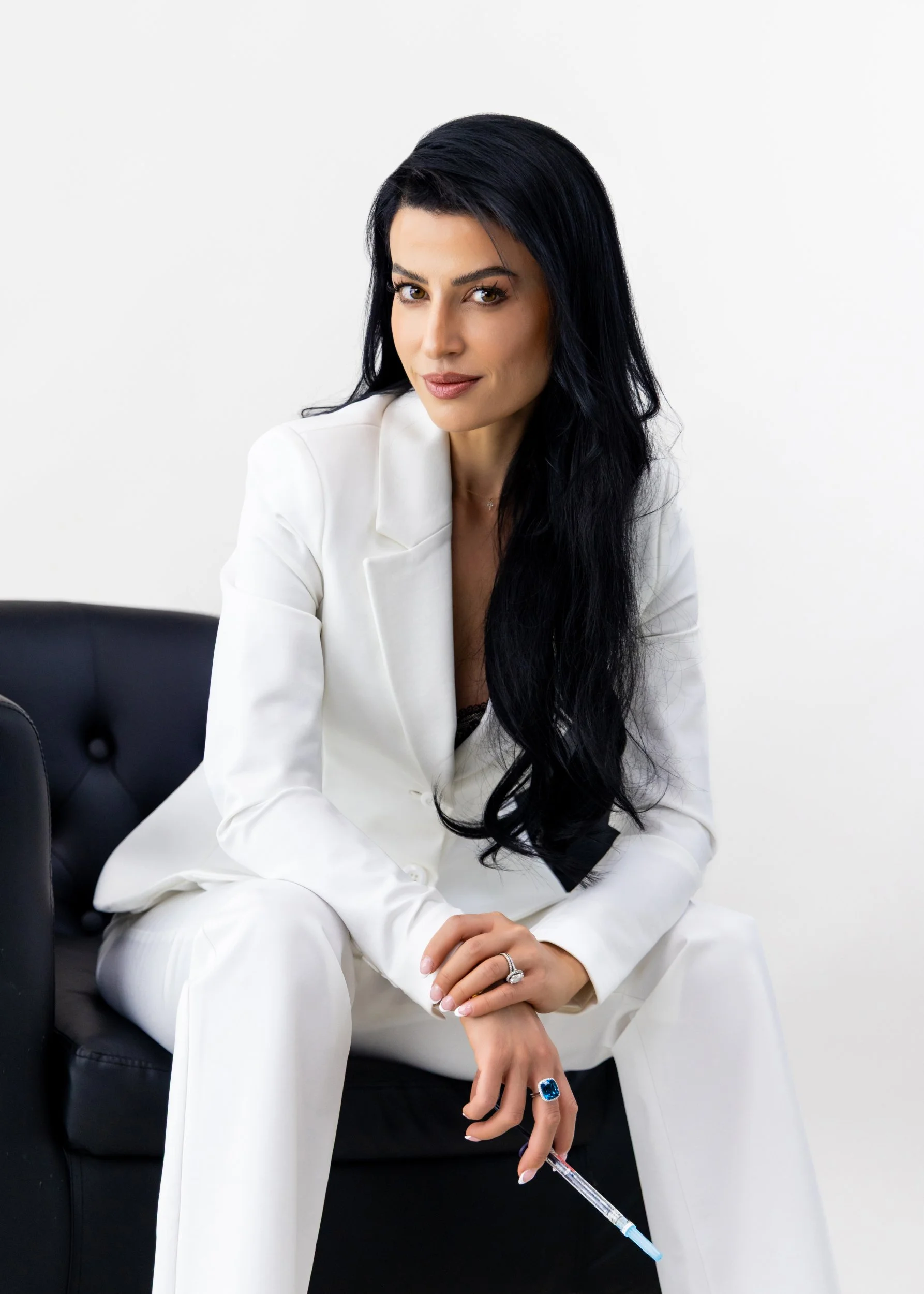 A woman with long black hair, wearing a white suit, sitting on a black chair, holding a syringe in her left hand, looking confidently at the camera against a plain white background.