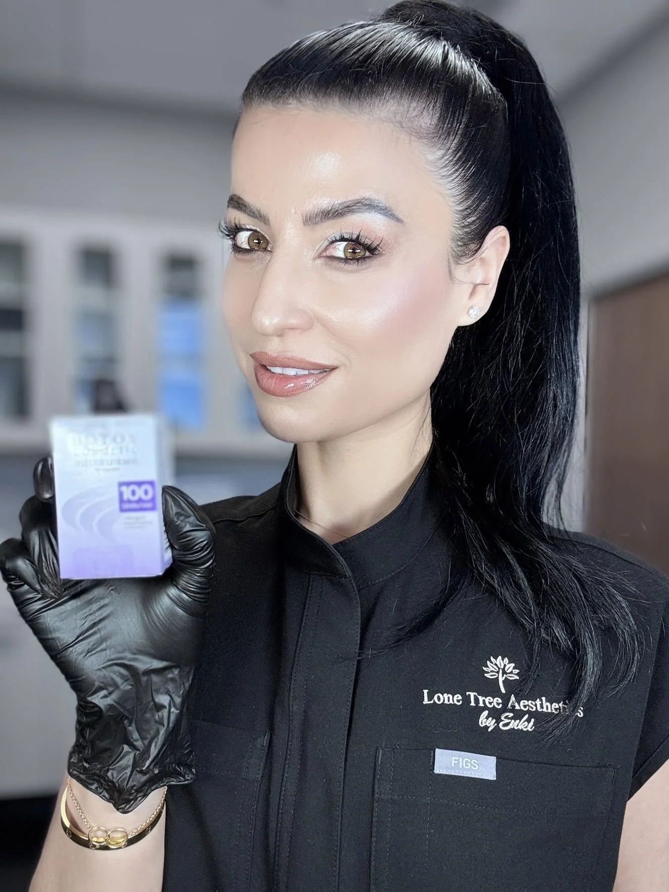 A woman with black hair and wearing black gloves holds a small box labeled '100'. She is smiling and wearing a black shirt with 'Lone Tree Aesthetics by Enki' embroidered on it.