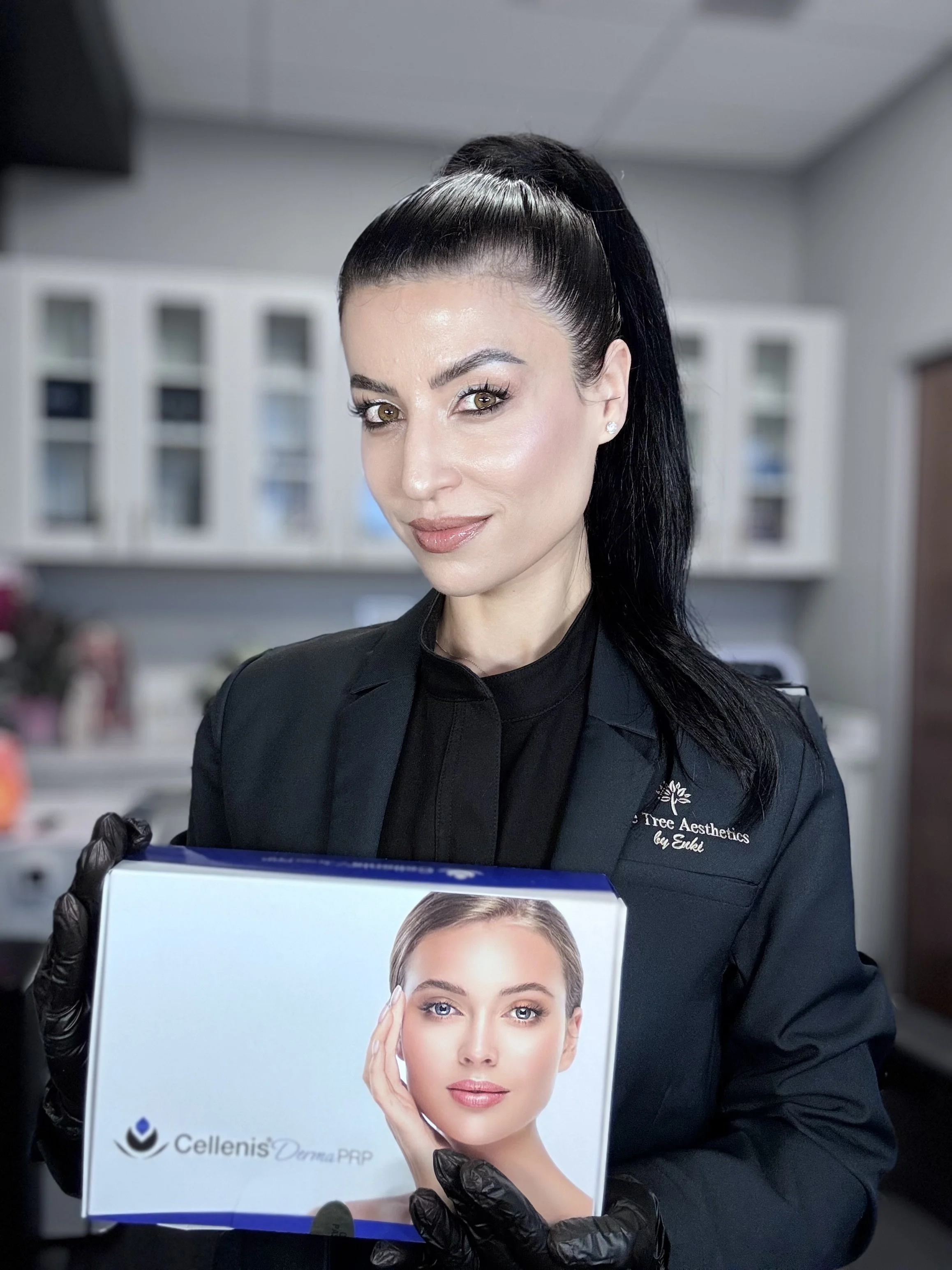 A woman with dark hair tied in a high ponytail, wearing a black blazer and gloves, holding a box with a woman's face on it. The setting appears to be an aesthetic clinic or office.