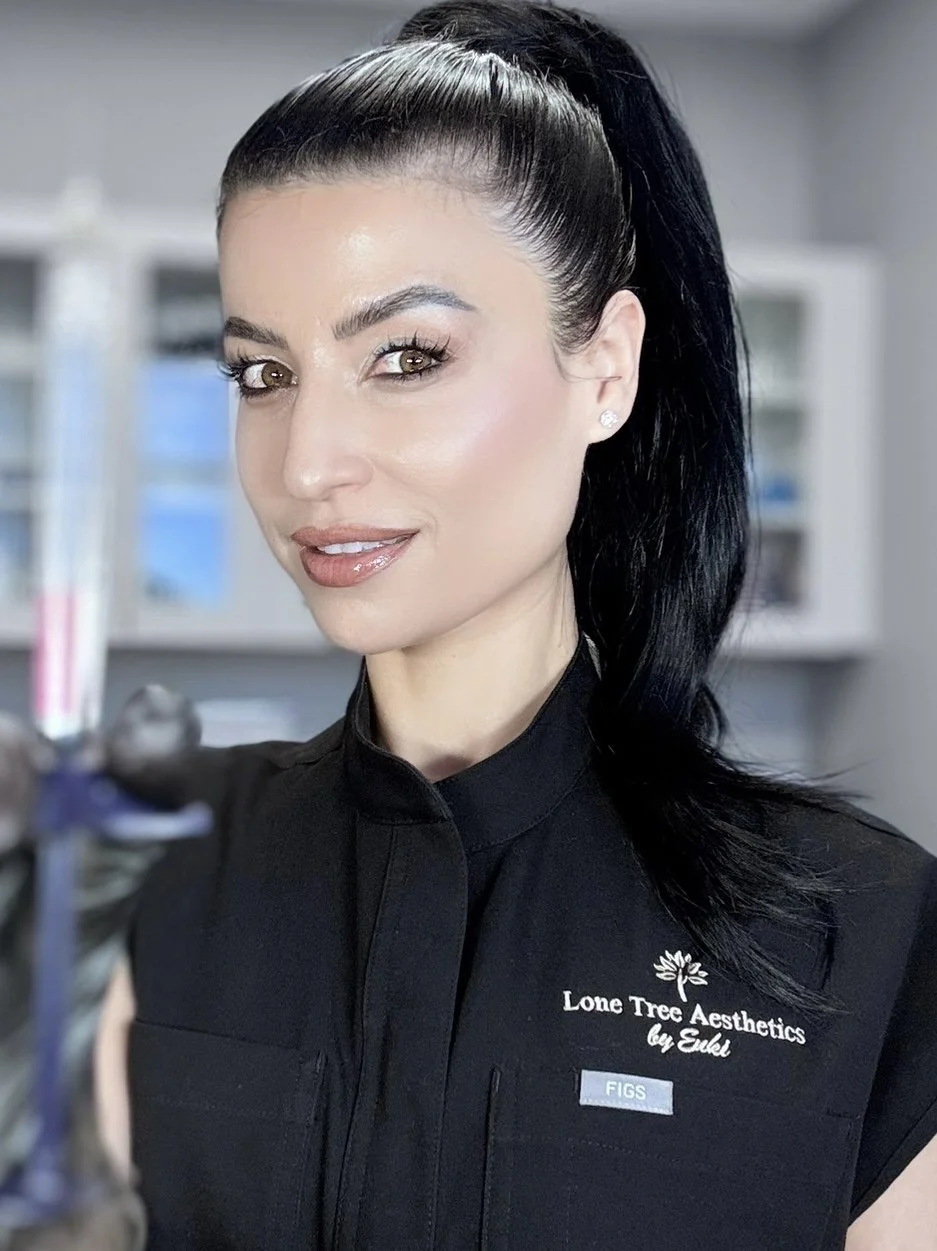 A woman with long dark hair tied back in a ponytail, wearing a black uniform with the logo 'Lone Tree Aesthetics by Eukl' and a name tag that reads 'FIGS,' is smiling in a clinical or medical setting.