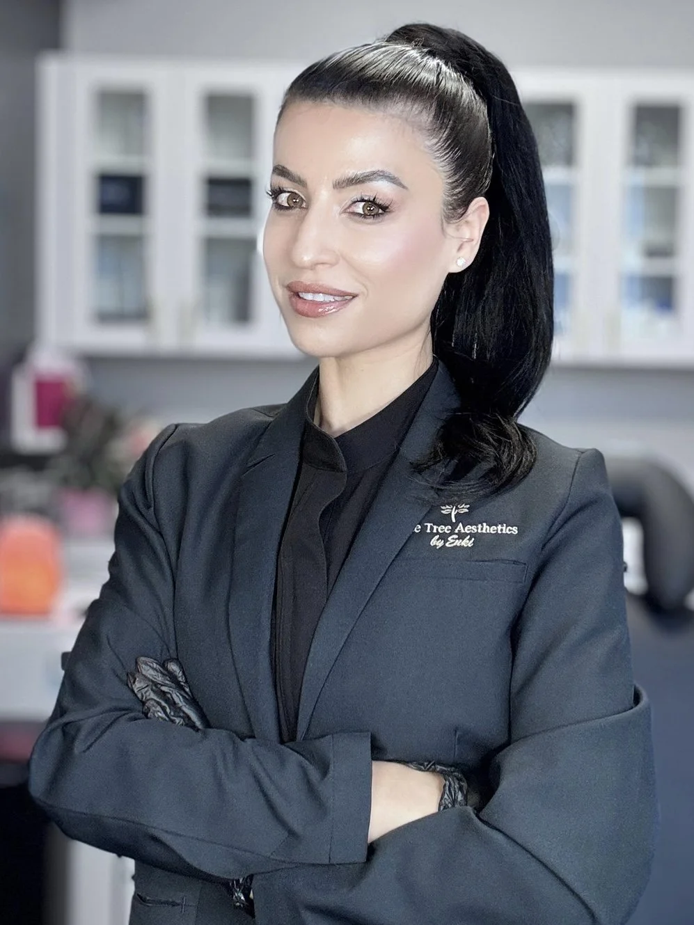 A woman with dark hair in a high ponytail, wearing a black blazer with embroidered text, smiling with arms crossed, in an office setting.