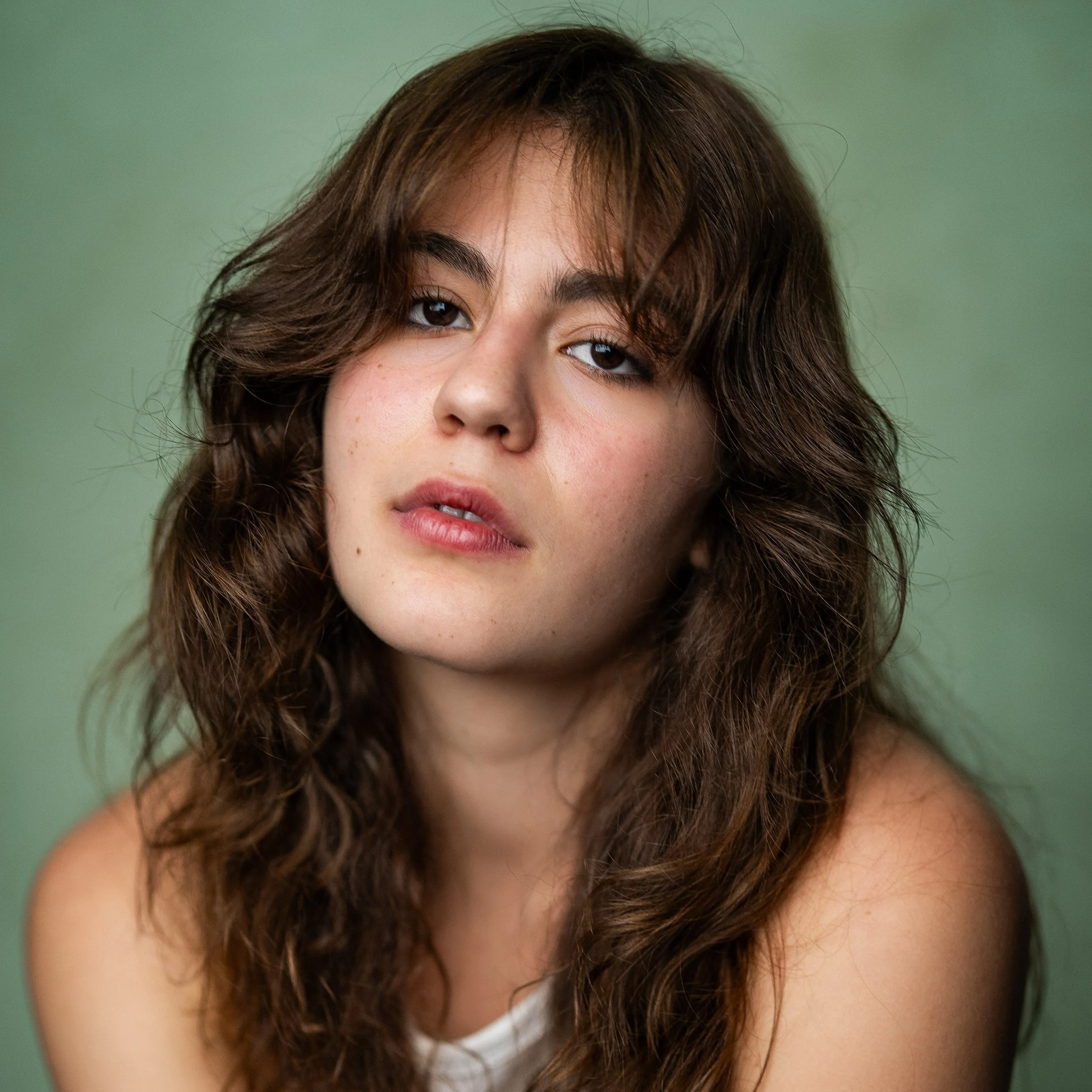 Close-up of a young woman with wavy brown hair and light skin against a muted green background.