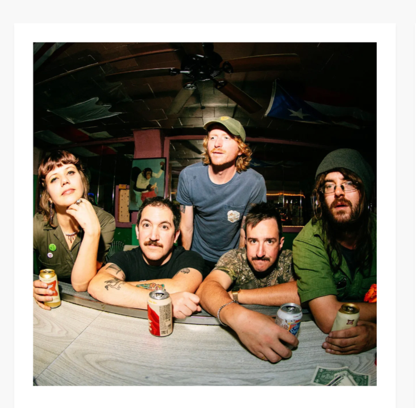 Five friends sitting at a bar counter with drinks in front of them, one person standing behind them, all inside a bar or pub with a ceiling fan and colorful decor.