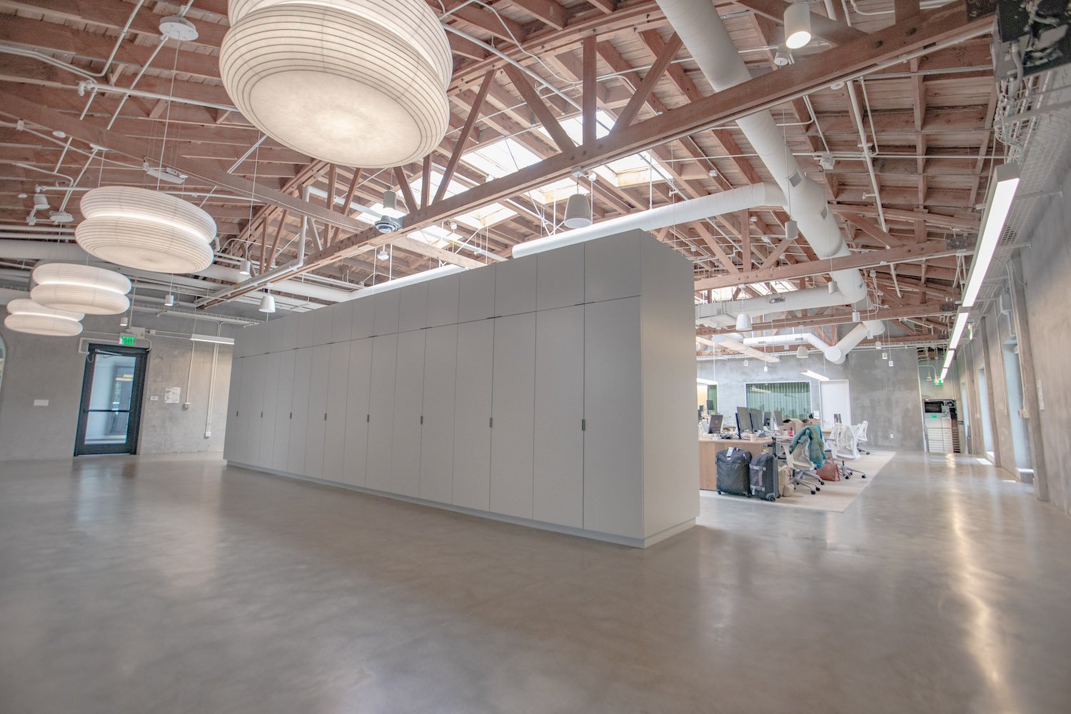 Modern office space with exposed ceiling, large hanging lights, white partition, and open seating area.