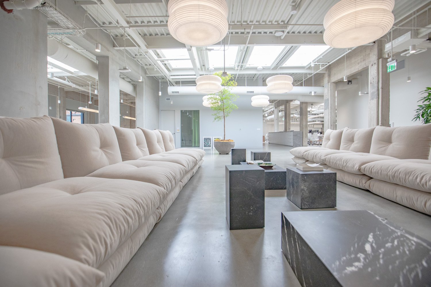 Modern office lobby with long beige couches, black marble coffee tables, overhead pendant lights, and a potted tree on a polished concrete floor.