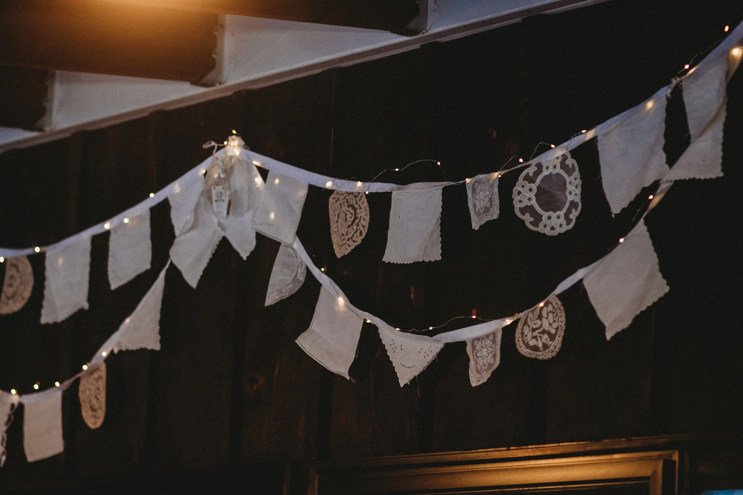 White fabric bunting with lace and embroidered patterns hanging from a ceiling, illuminated by small string lights in a dark room.