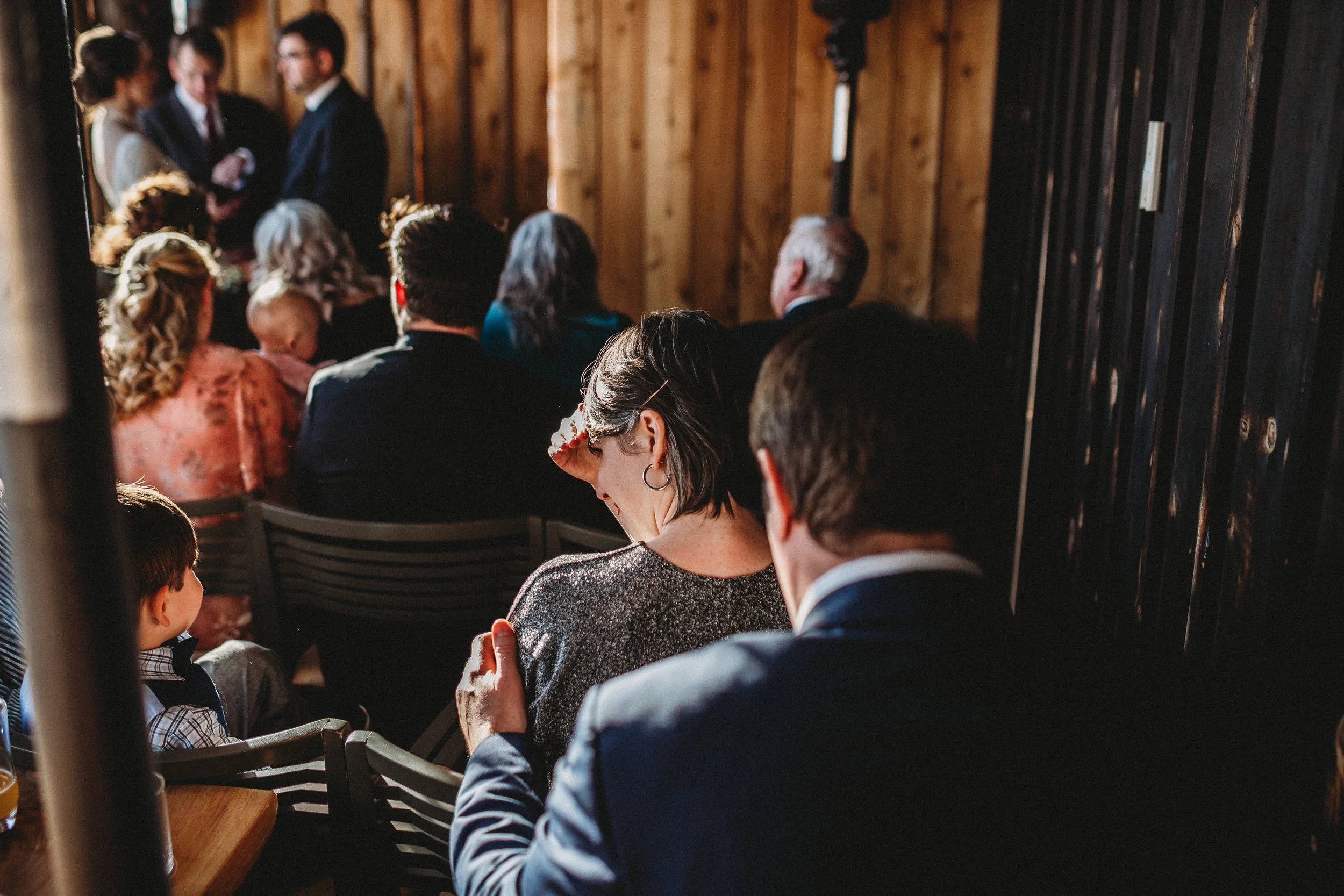 People attending a wedding ceremony inside a rustic wooden venue, some seated and others standing at the altar.