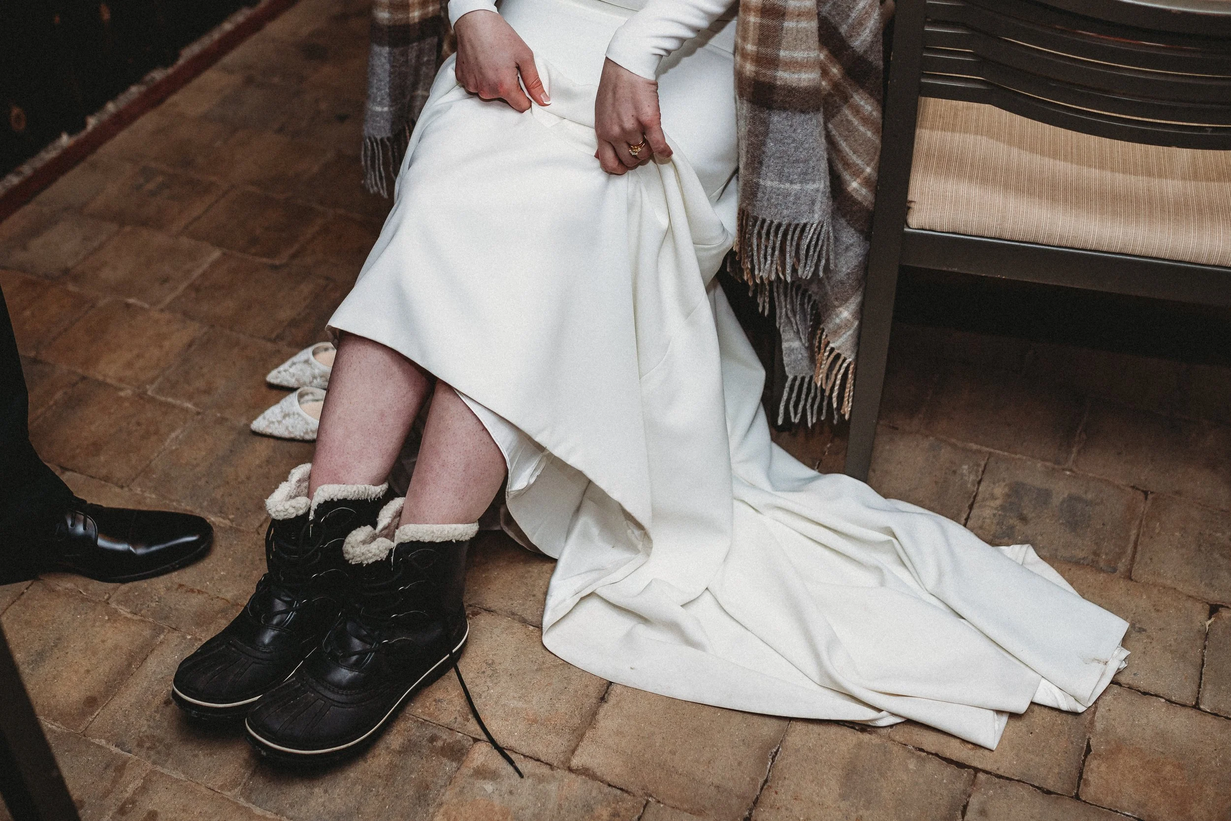 Person in a white wedding dress with black boots sitting on the floor, lifting the dress to reveal their legs and shoes, with a plaid blanket draped over their lap, next to a stack of chairs.