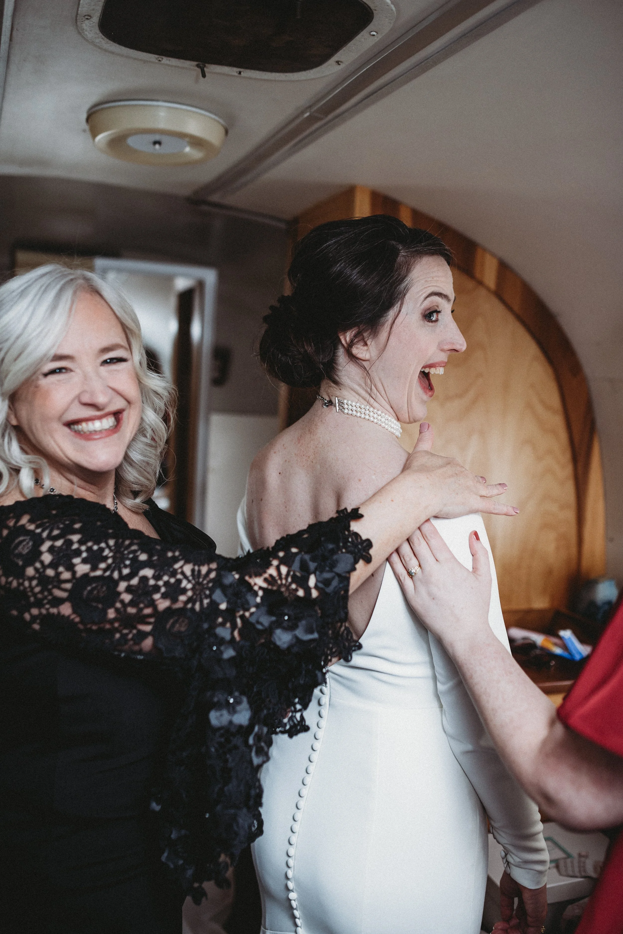 A bride with dark hair and a pearl necklace, smiling and playfully posing while getting ready for her wedding, with a woman in black lace dress smiling beside her.