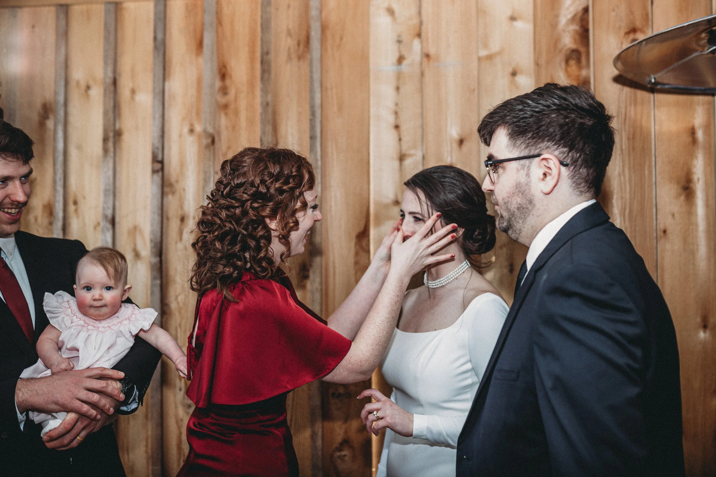 A woman with red curly hair is touching the face of a woman in a white dress, possibly during a wedding, while a man in a dark suit stands nearby. A man holding a baby is on the left, all in front of a wooden wall.