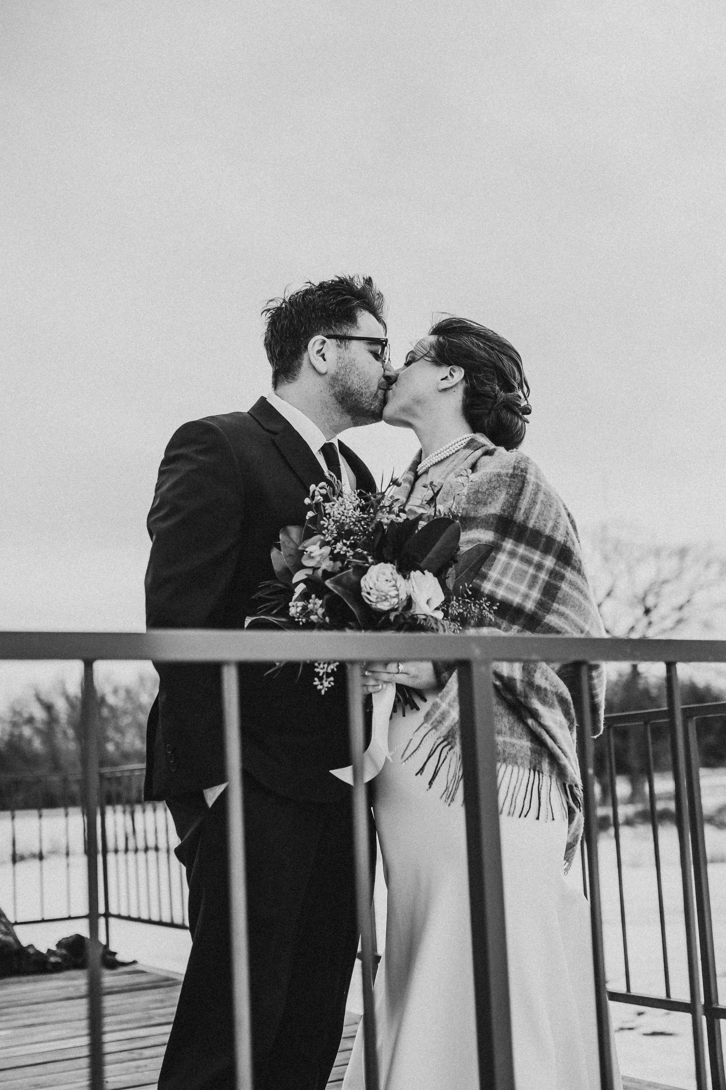 A black and white photo of a couple kissing on a bridge, with the woman holding a bouquet of flowers. The man is wearing a suit and tie, and the woman is wrapped in a plaid shawl.