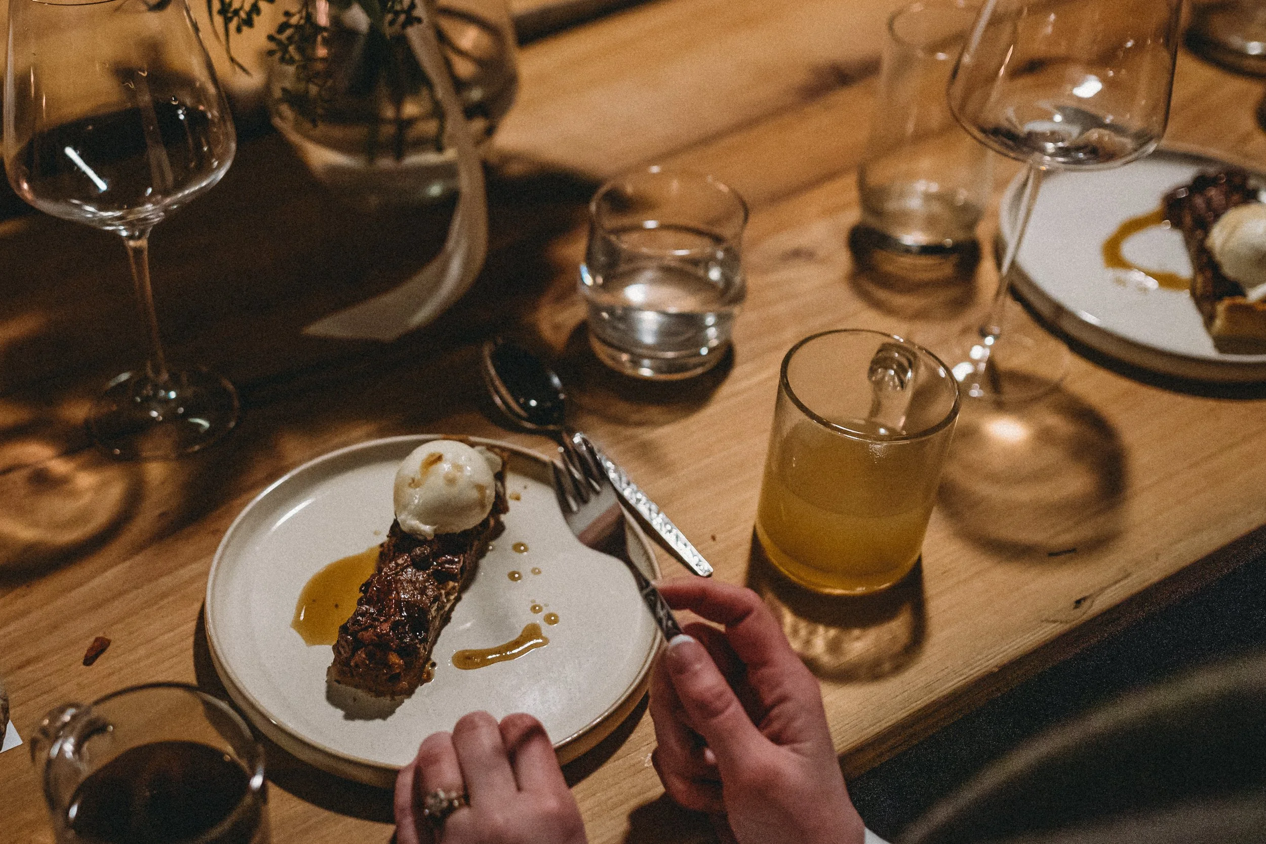 A dessert on a white plate with a scoop of ice cream, chocolate topping, and caramel sauce, on a rustic wooden table surrounded by glasses of water, wine, and a yellow beverage, with a person's hands holding a fork and knife.