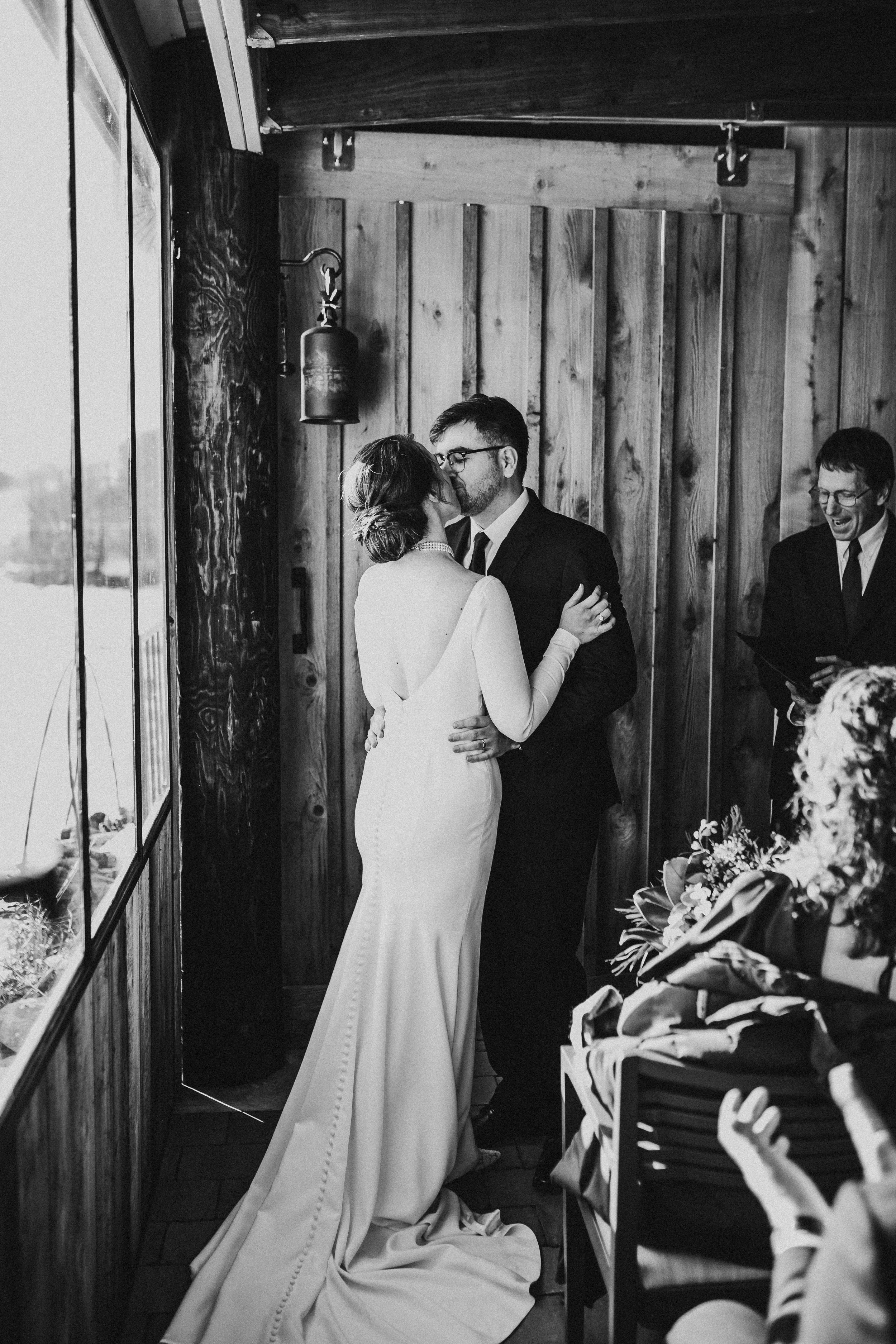 A black and white photo of a wedding ceremony where a couple is kissing, the bride wearing a long gown and the groom in a dark suit, inside a wooden venue near large windows.