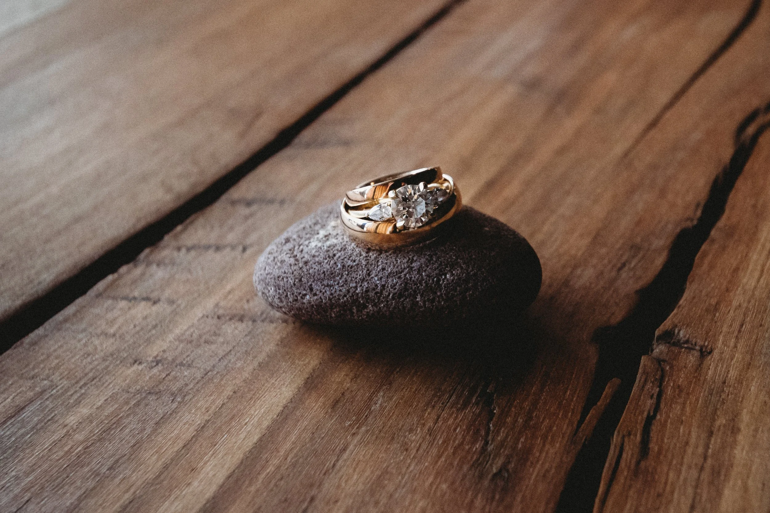 A gold ring with a large central diamond and smaller surrounding diamonds resting on a small dark stone placed on a wooden surface.
