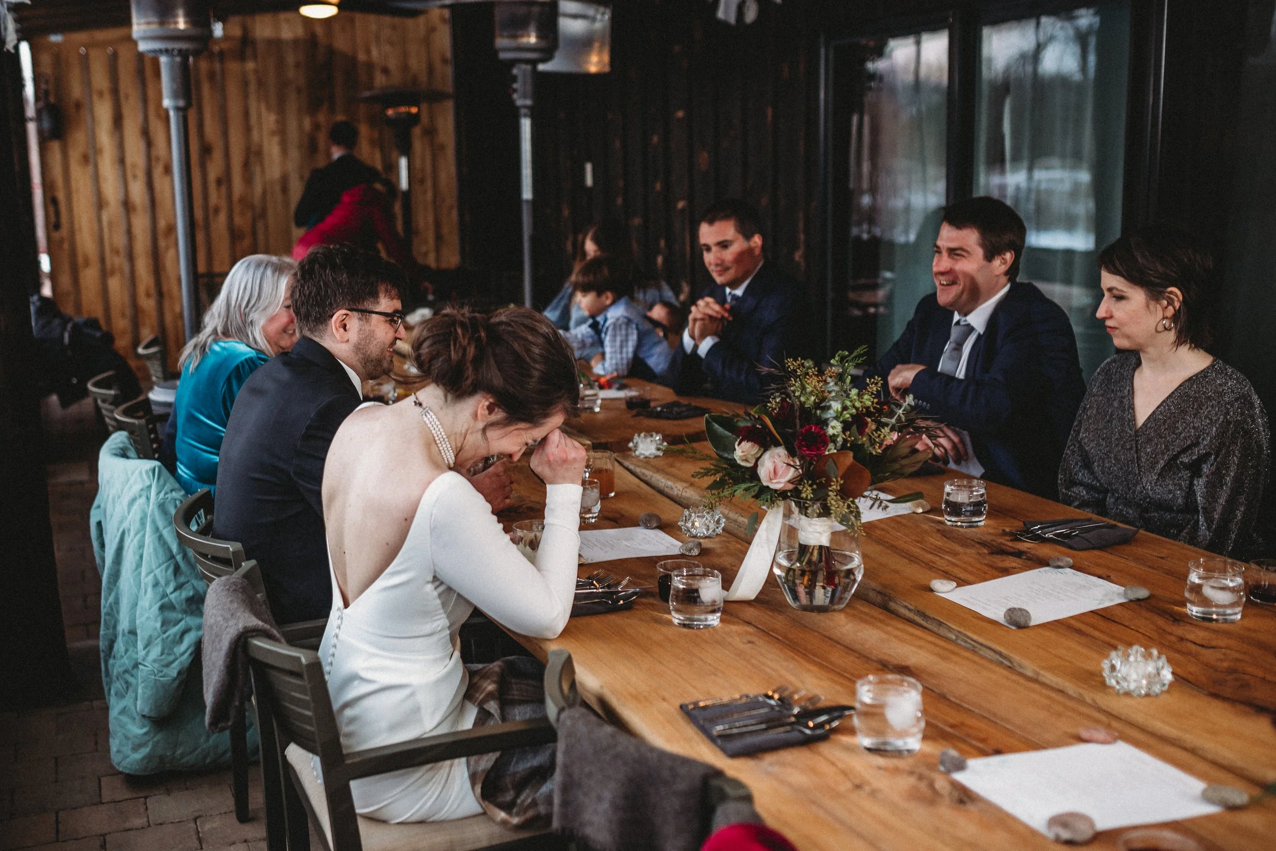 A group of people sitting at a long wooden table during a celebration, with a woman in a white dress appearing emotional and crying.