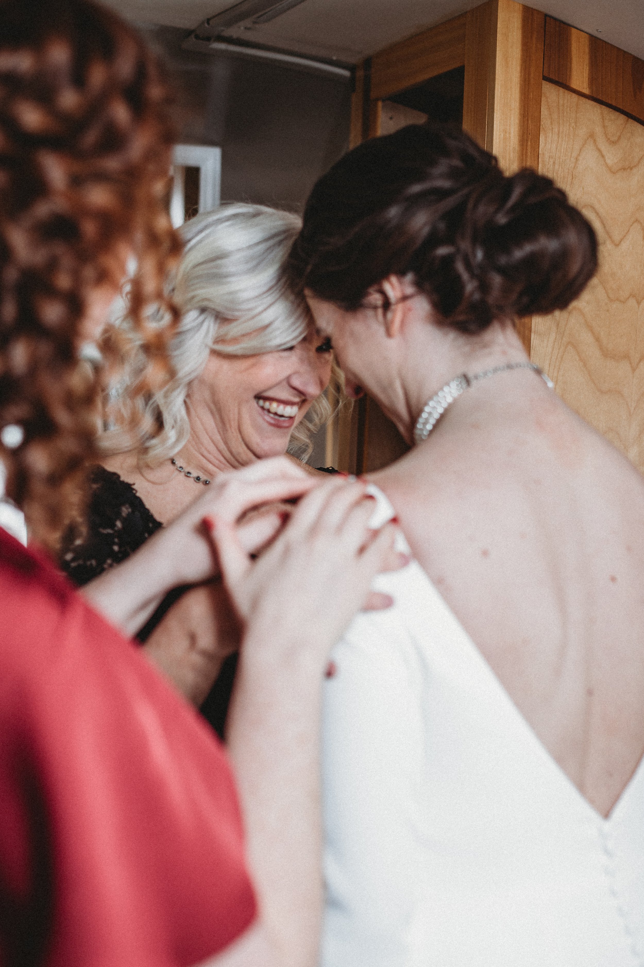 Two women sharing an intimate, joyful moment, possibly a bride and her mother, during a wedding or celebration, with one woman in a white dress and the other in a black dress, both smiling and leaning close.
