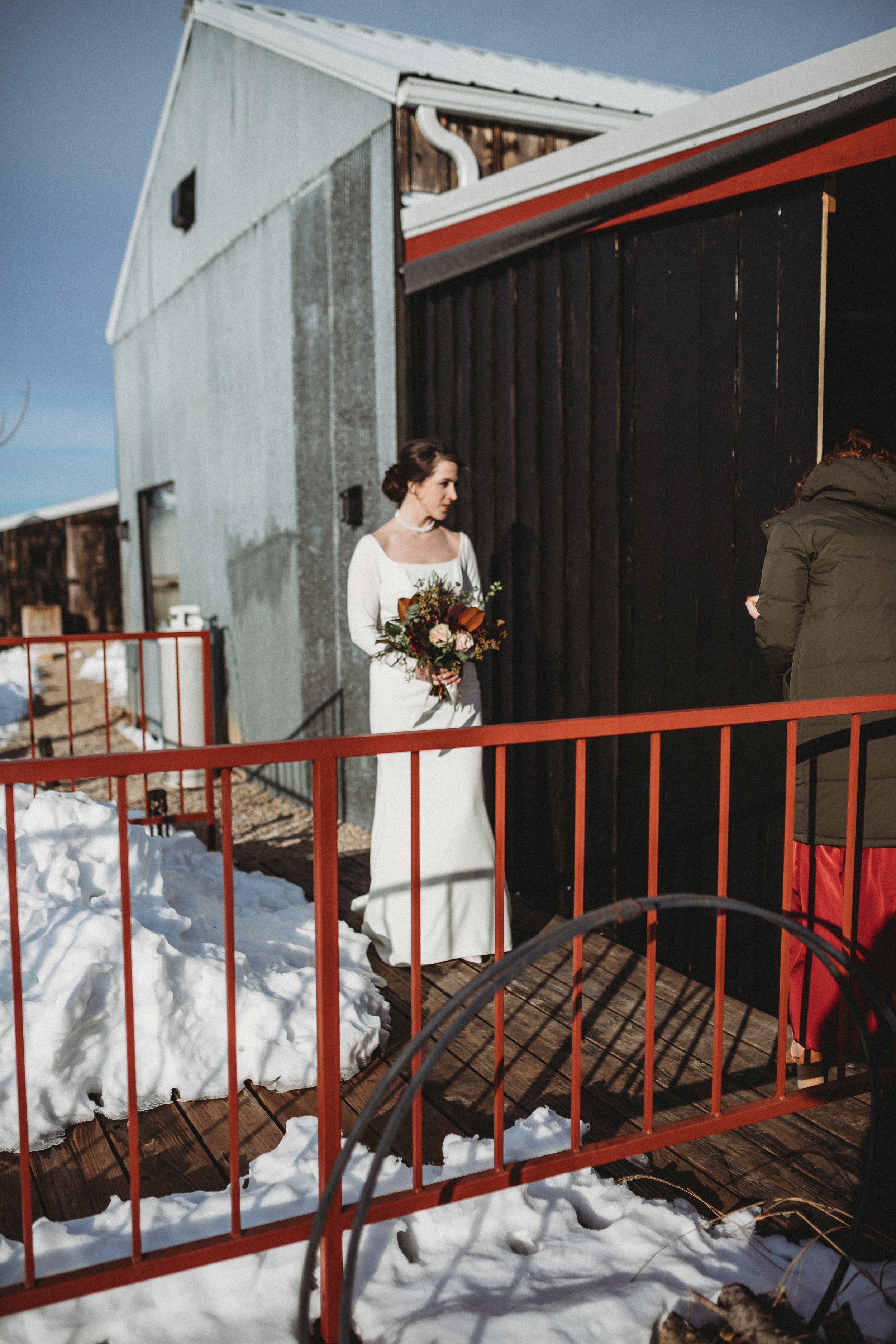 A woman in a white wedding dress holding a bouquet of flowers standing outdoors on a wooden deck, near a black building, with snow on the ground and a clear blue sky.