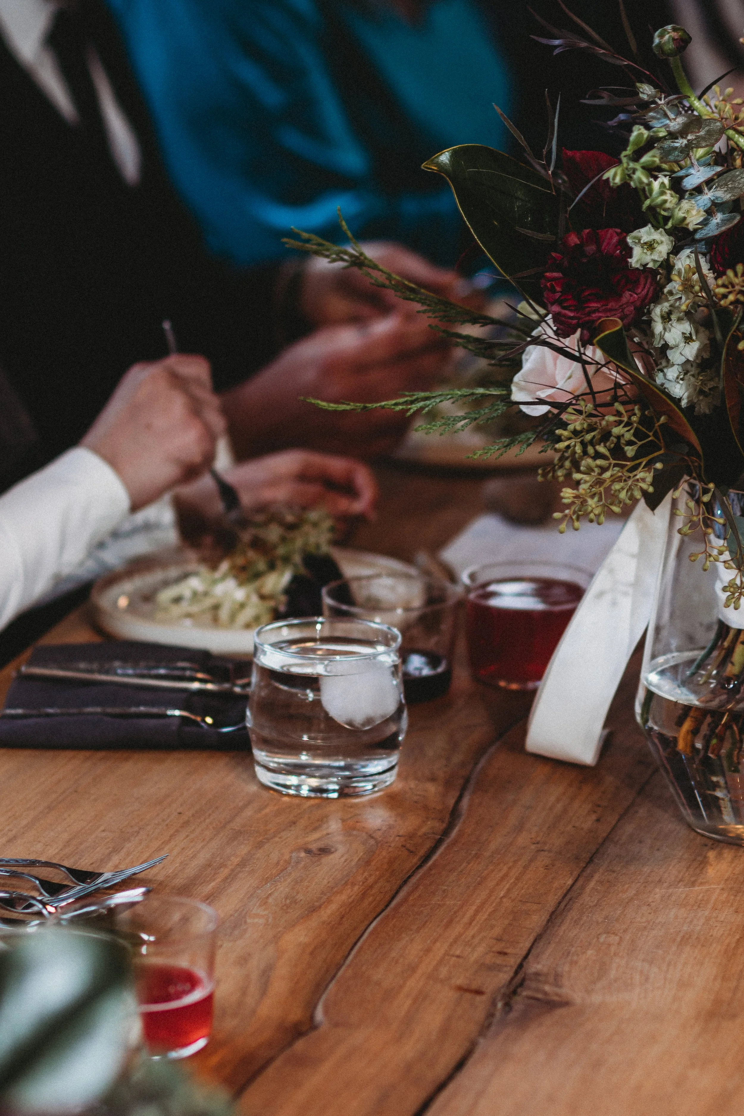 People dining at a wooden table with a floral centerpiece, glasses of water, and plates of food.
