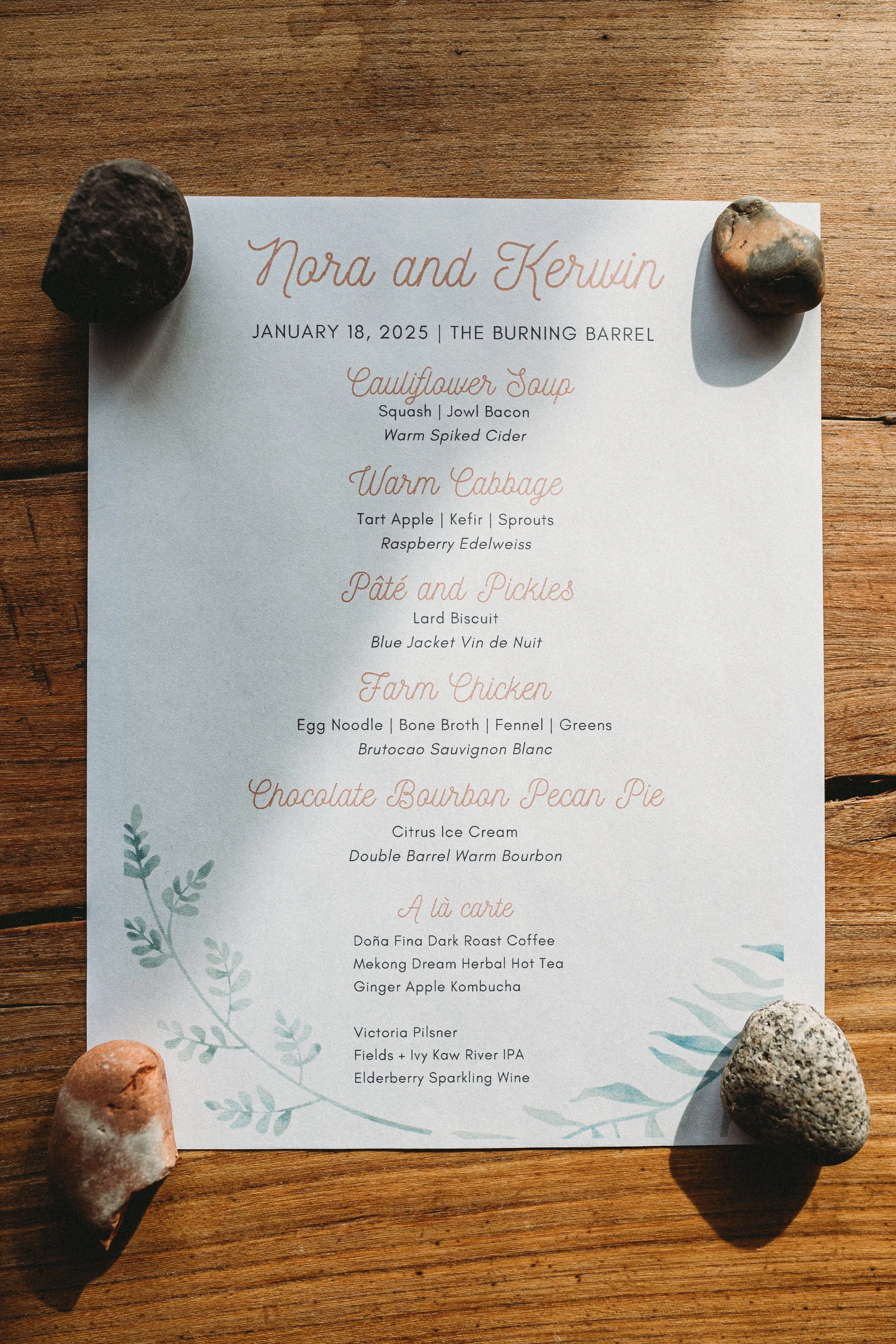 Wedding menu on white paper with decorative leaves, held down by four stones on a wooden surface.