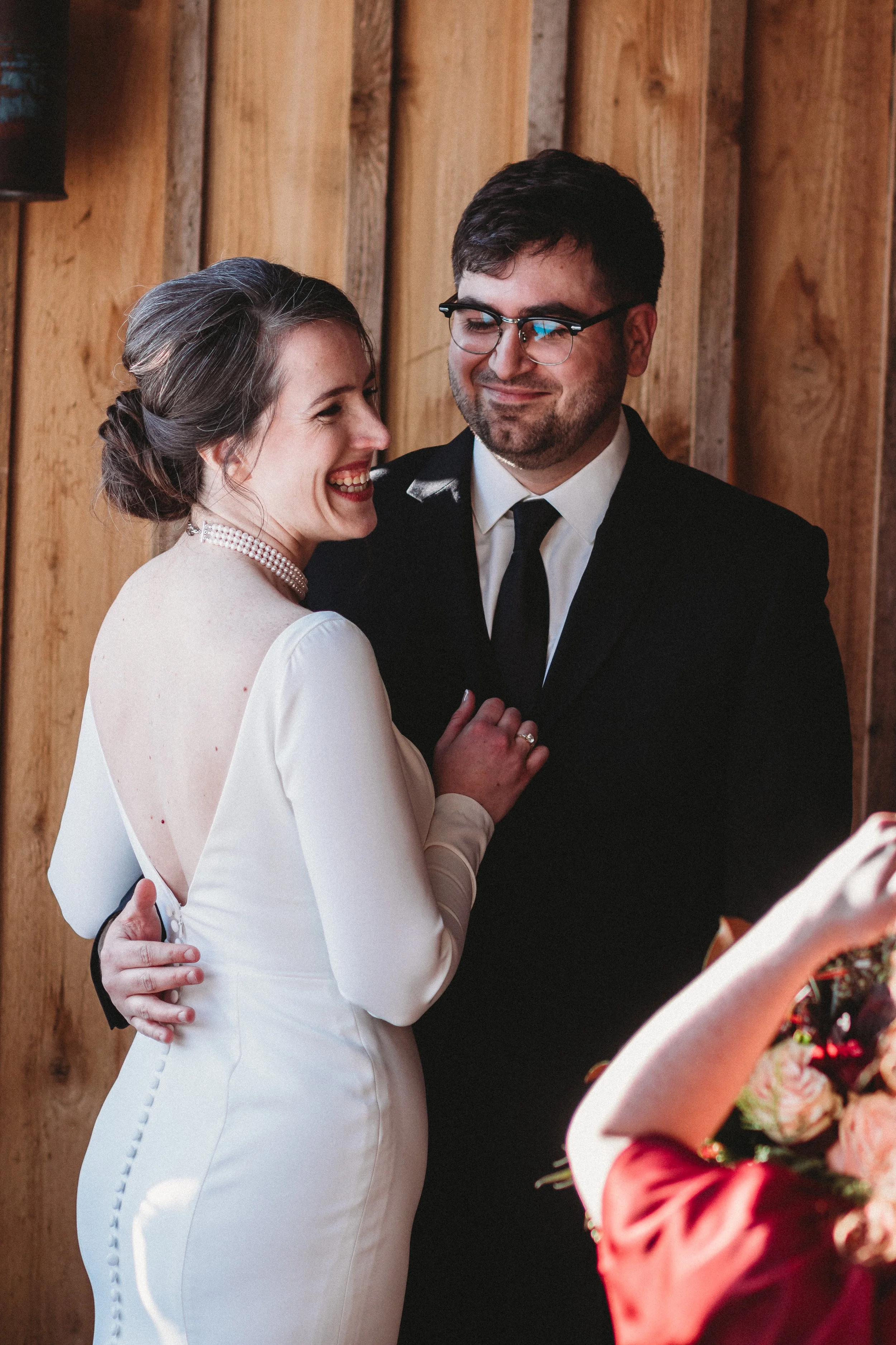 A bride and groom sharing a happy moment, with the bride smiling and the groom smiling gently, indoors with wooden paneling in the background.