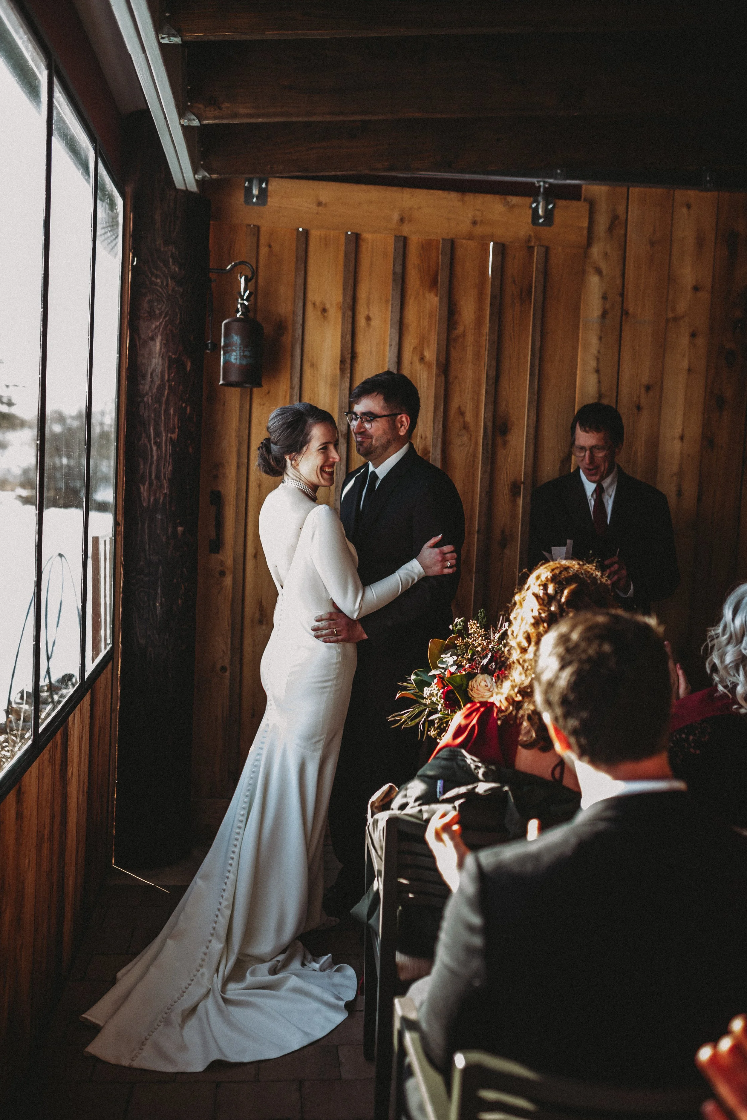 Bride and groom sharing their wedding vows indoors, with wedding guests seated nearby, wooden walls in the background, and sunlight coming through a window.