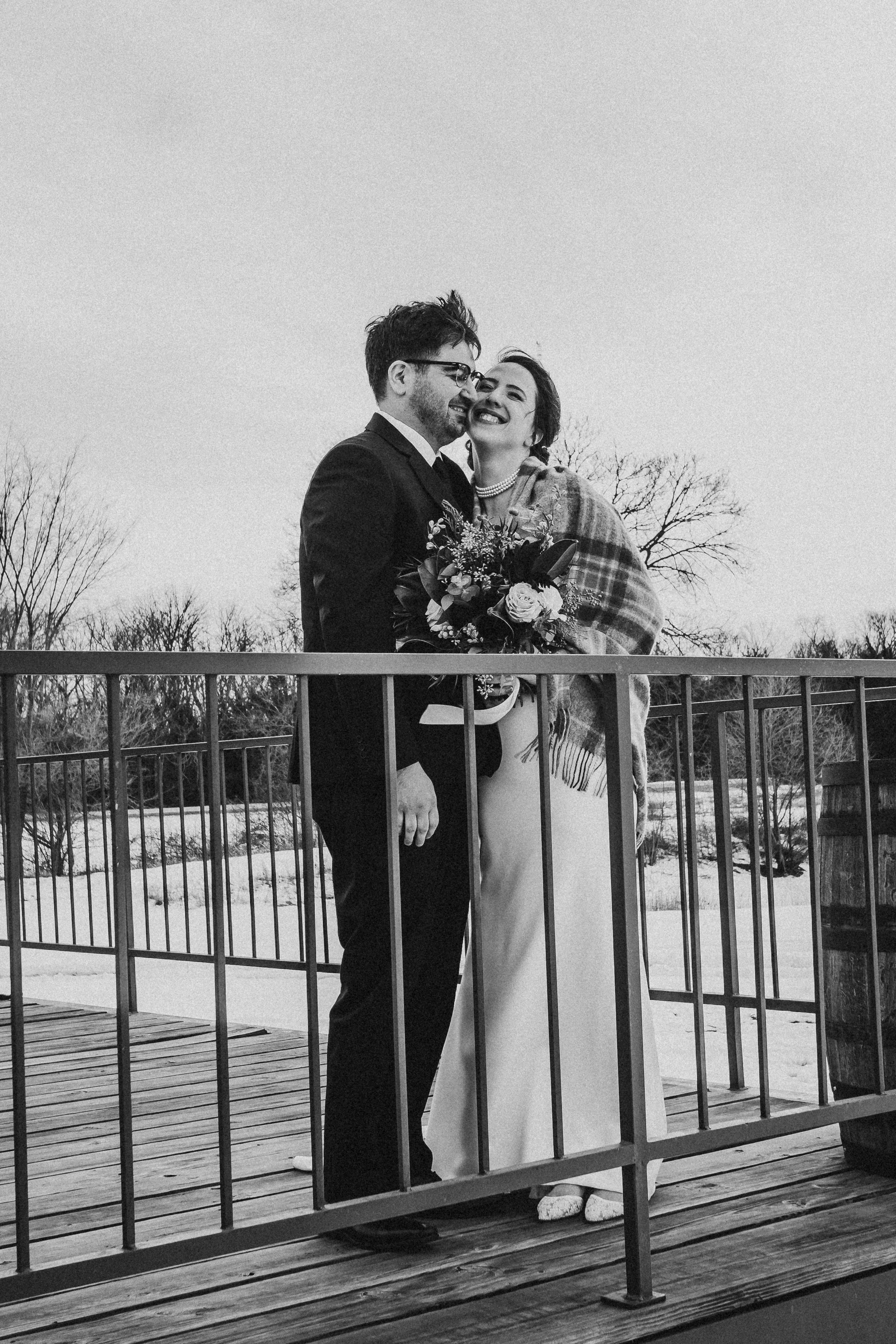 Black and white photo of a bride and groom smiling together on a wooden balcony, with a snowy landscape and leafless trees in the background.