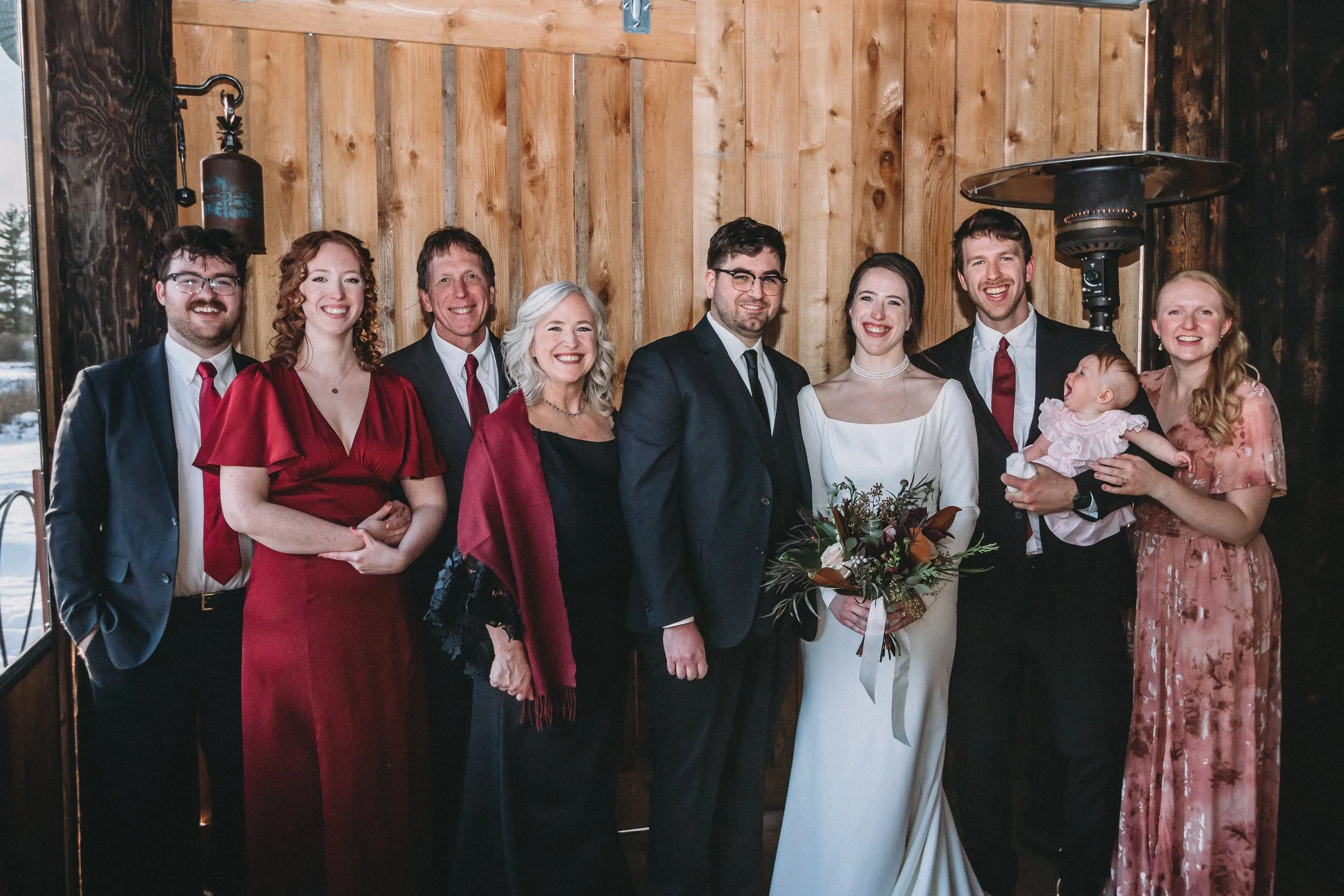 Group of nine people dressed in formal attire, including a bride and groom, standing indoors against a wooden wall, smiling at the camera.