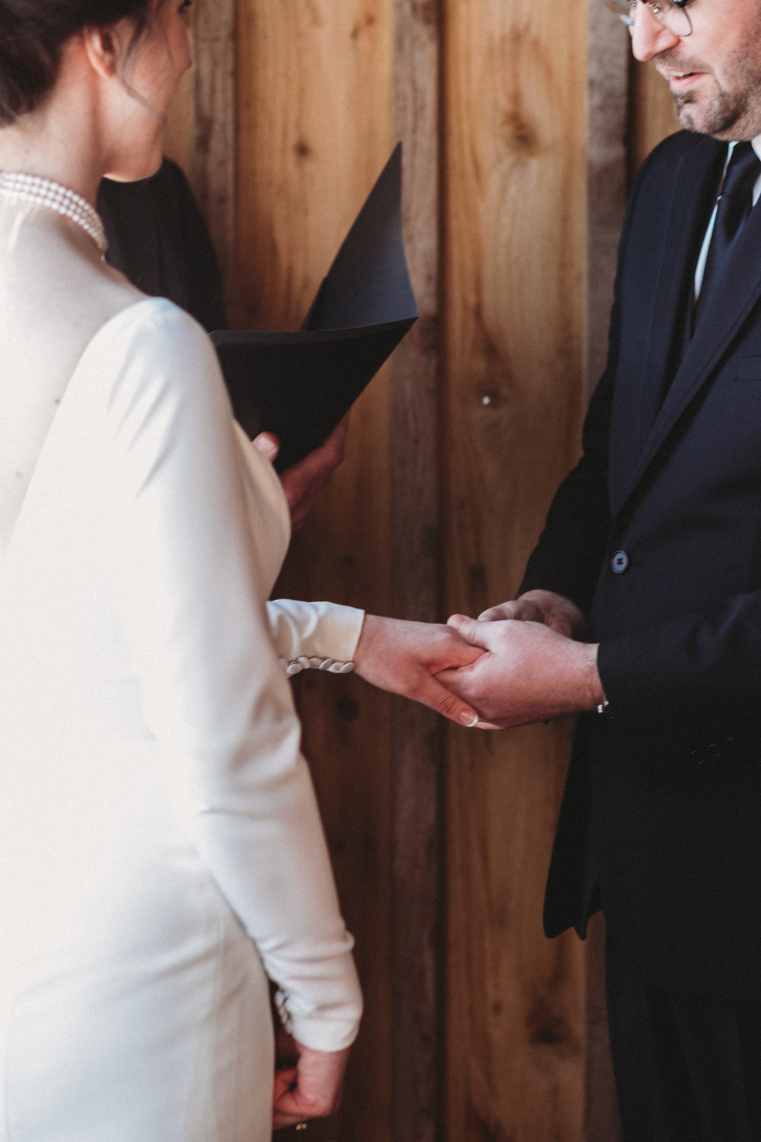 A wedding ceremony with a bride and groom holding hands, exchanging vows, at a venue with wooden walls.