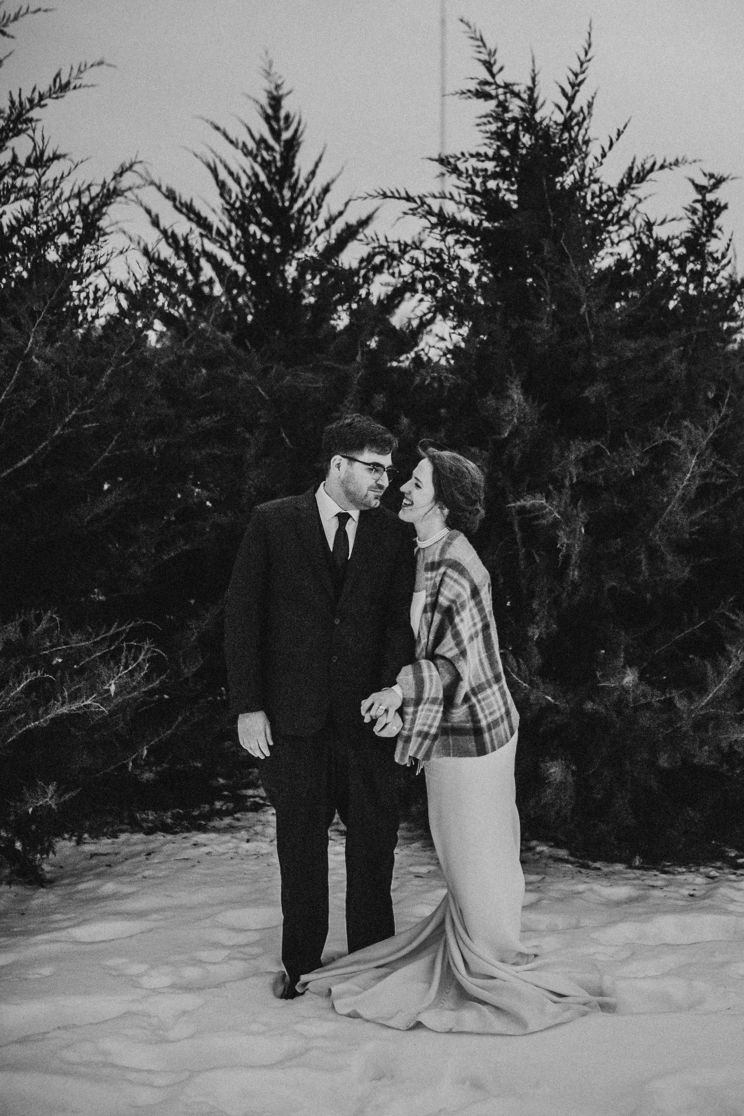 A black-and-white photo of a man and woman holding hands, standing in snow with pine trees in the background.