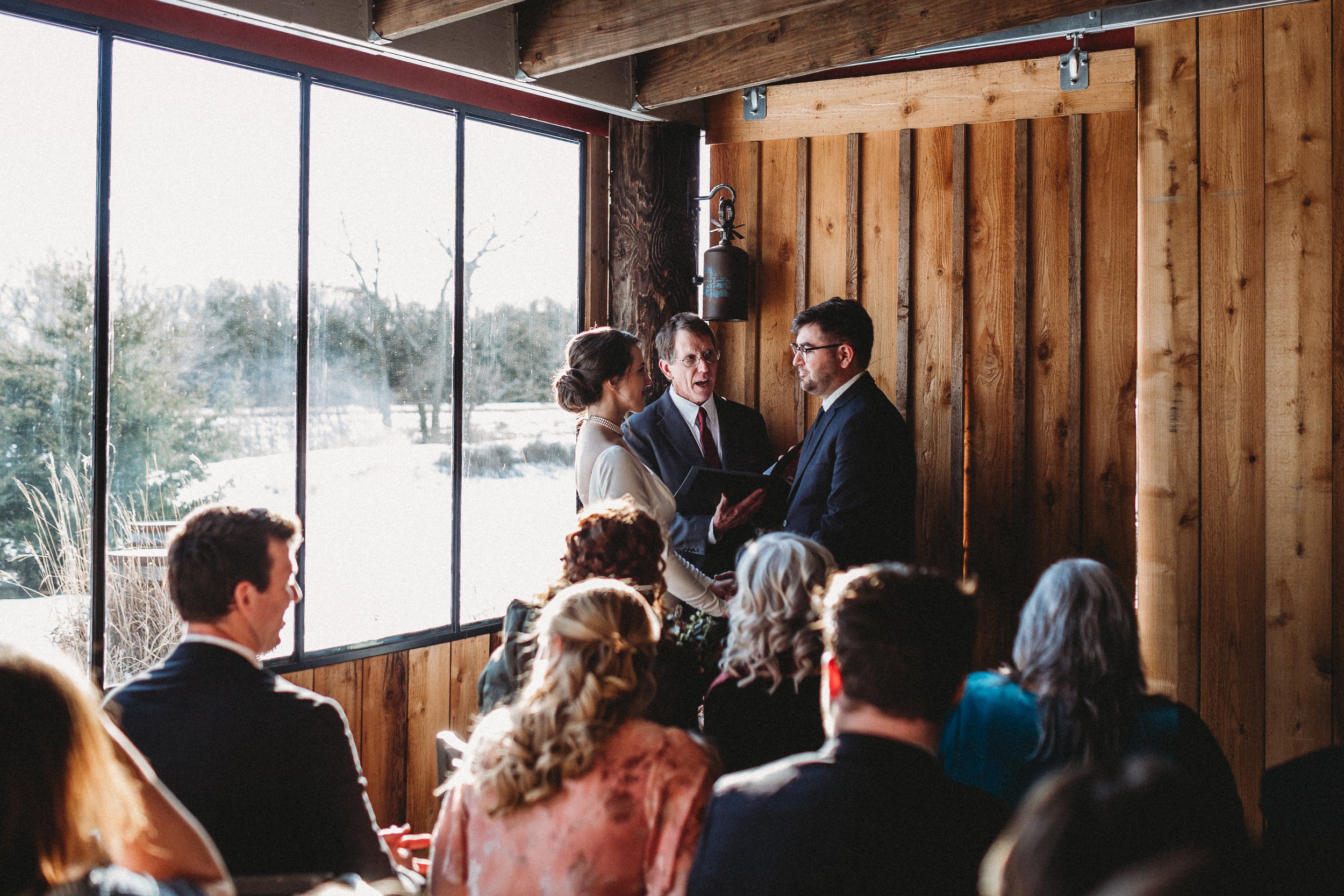 A couple getting married during a wedding ceremony, with an officiant officiating in front of a large window showing a snowy landscape outside. Guests are seated and watching the ceremony.