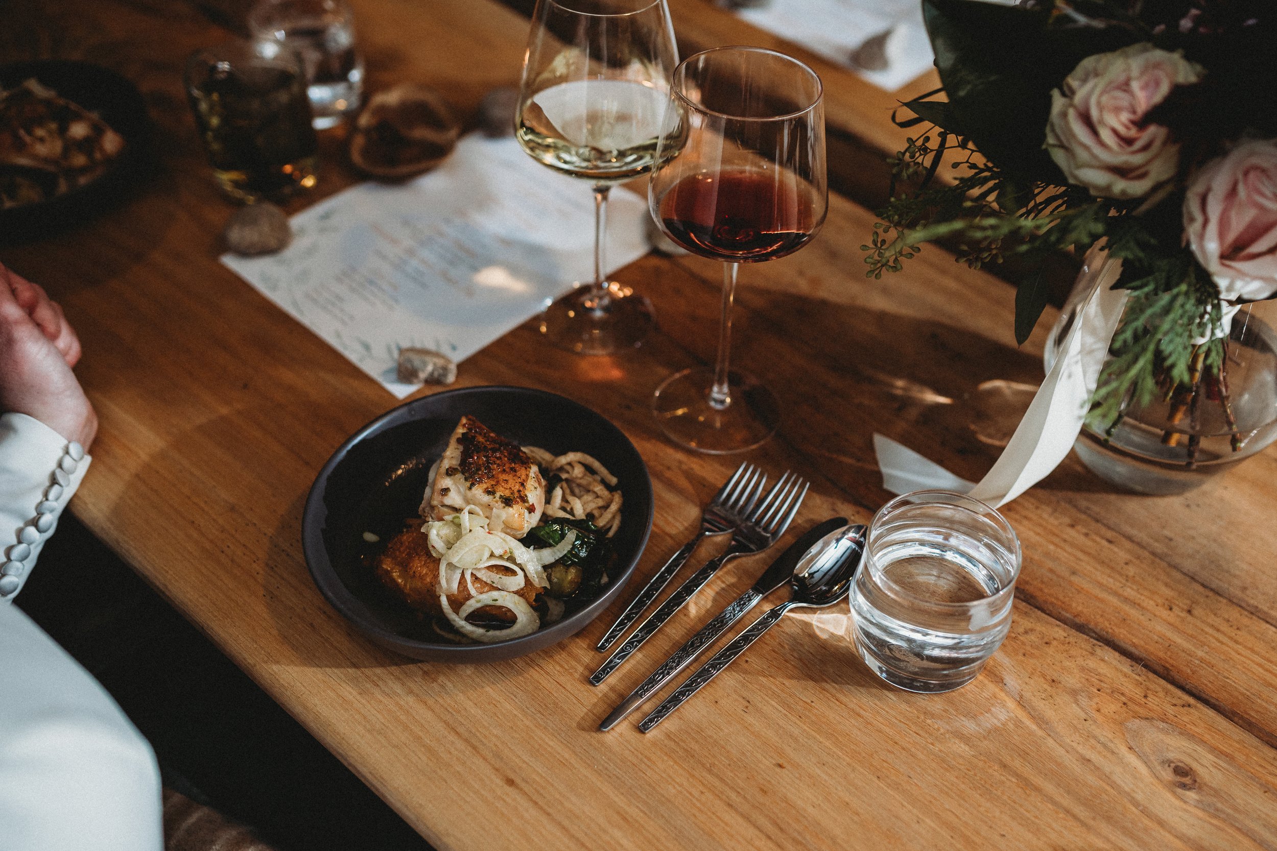 A dinner table set with a black plate of grilled chicken with onions and noodles, two glasses of white and red wine, a glass of water, cutlery, a menu, and a vase of pink roses on a wooden table. Burning Barrel Kansas