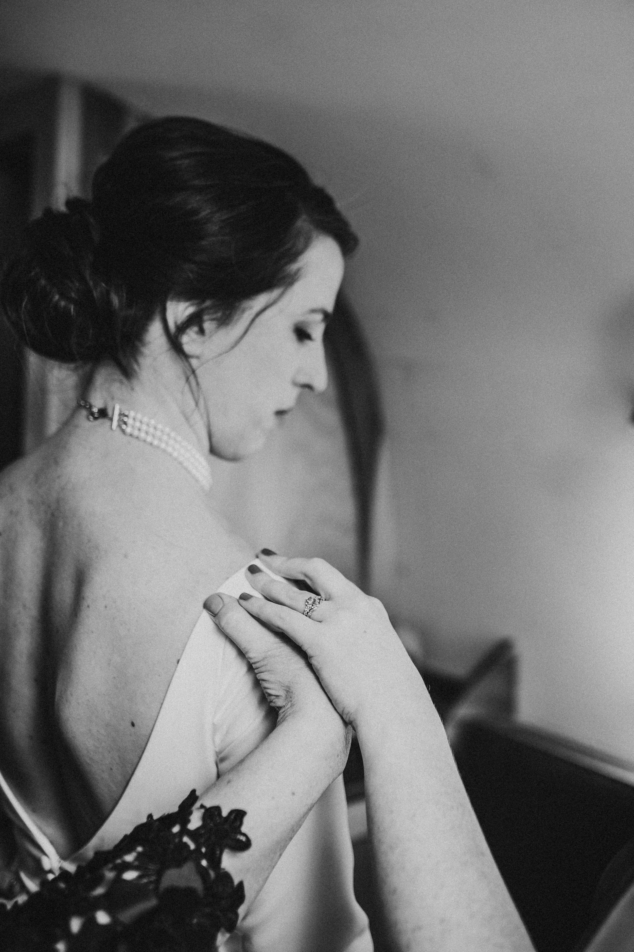 A black-and-white photo of a woman with dark hair styled in an updo, wearing a pearl necklace, and a wedding ring on her finger, showing her shoulder while a person adjusts her dress.