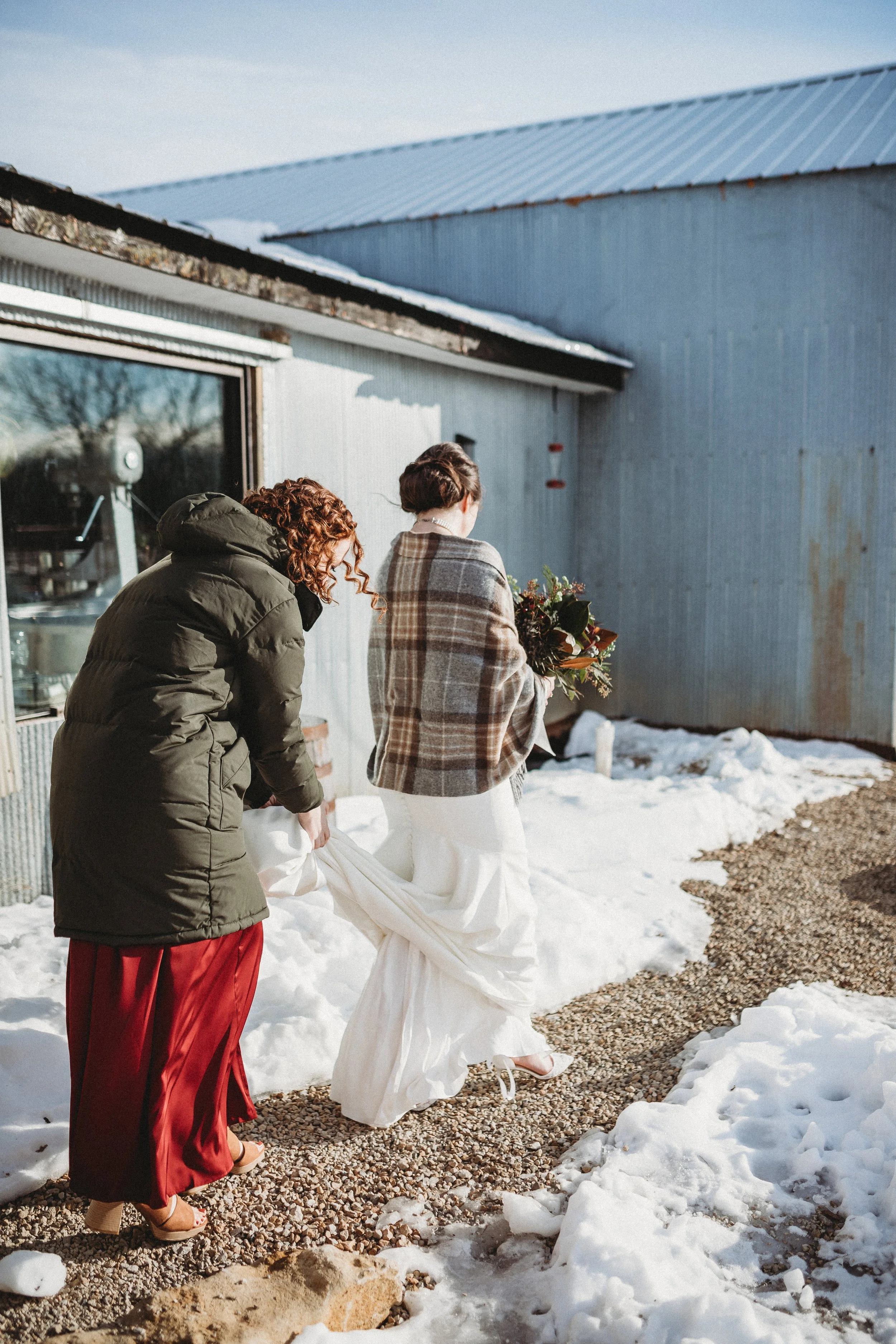 A bride in a white wedding dress holding a bouquet of flowers, walking outside on a snowy day, with one woman helping her with her dress.