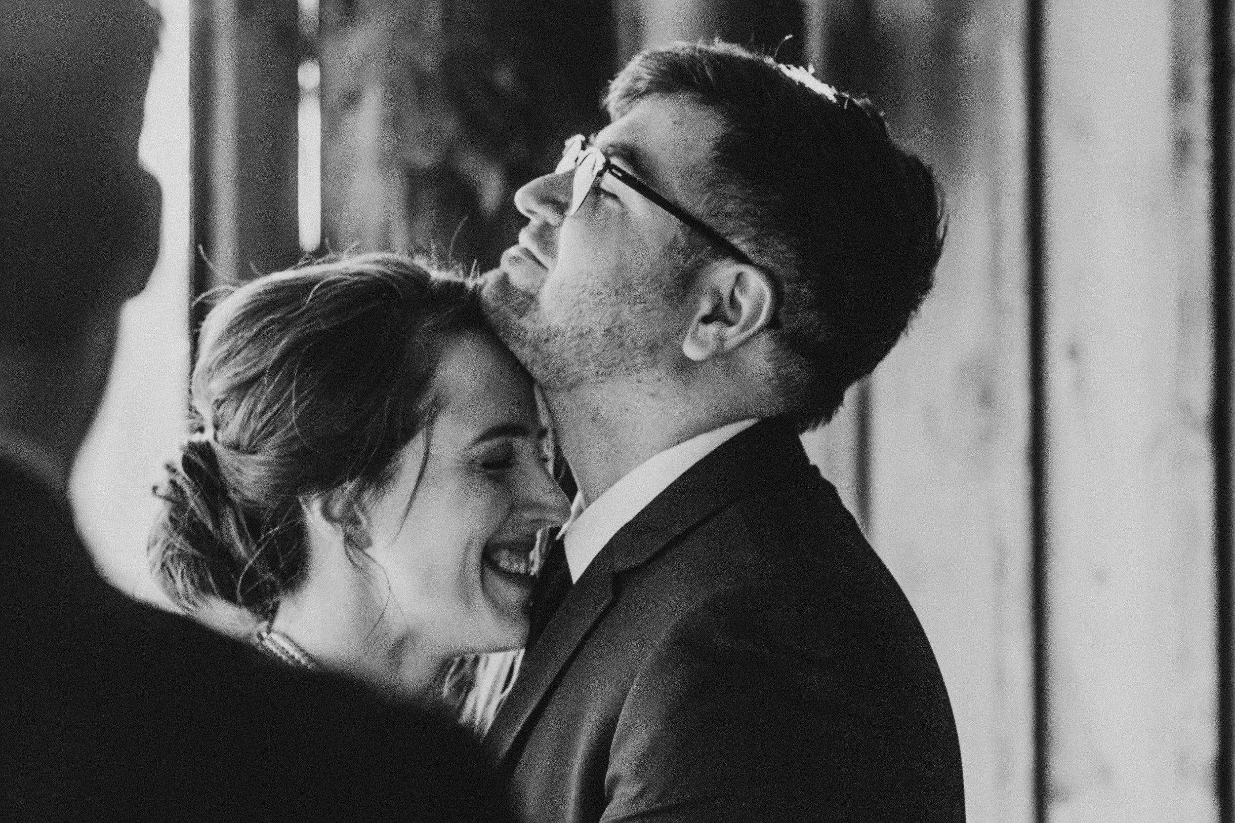 Black and white photo of a woman and a man in a wedding ceremony, with the woman smiling and the man kissing her on the forehead.