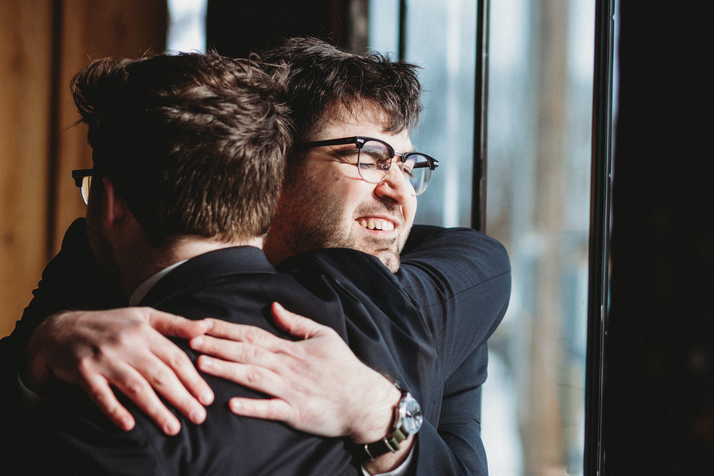 Two men embracing warmly, one with glasses and a beard smiling as they hug near a window.