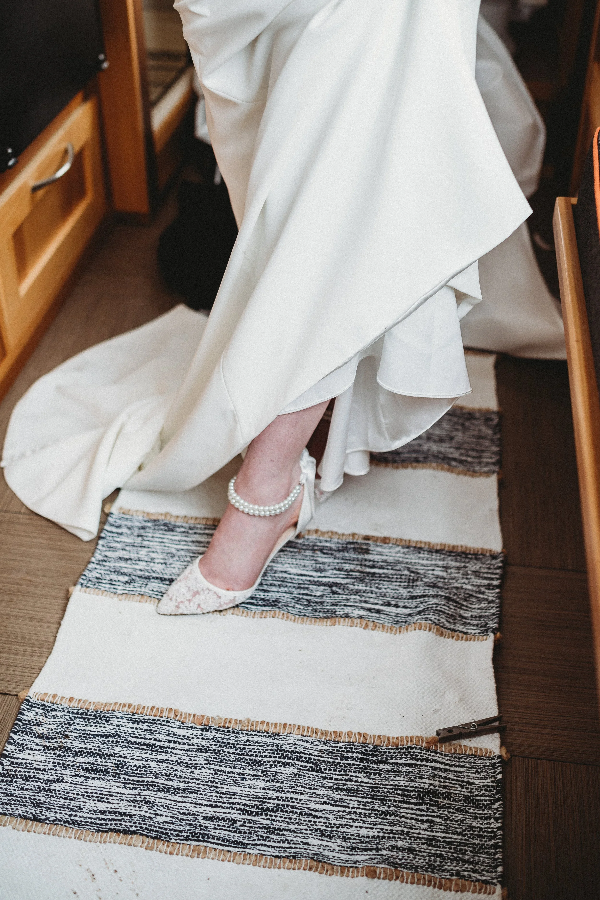 A bride wearing a wedding gown and pearl-anklet shoes, standing on a woven rug.